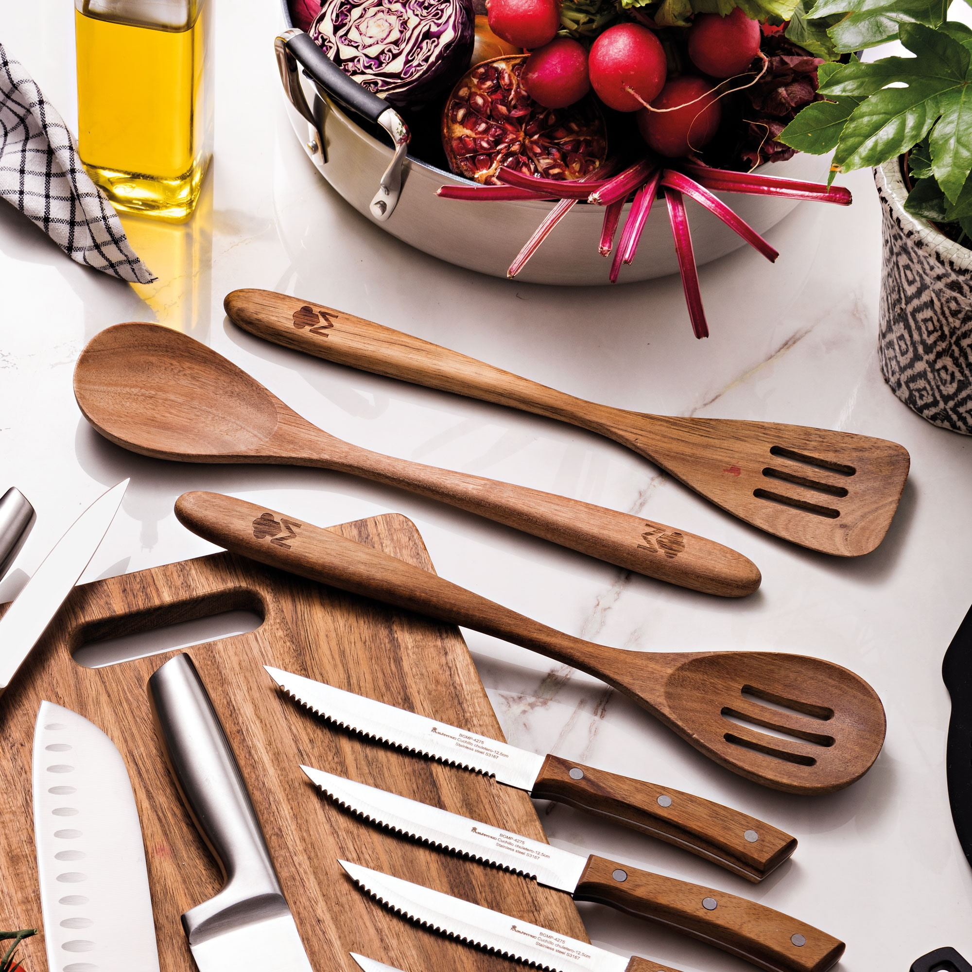 Cucharas de madera y cuchillos sobre una tabla de cortar, verduras en un cuenco y una botella de aceite sobre una superficie de mármol.