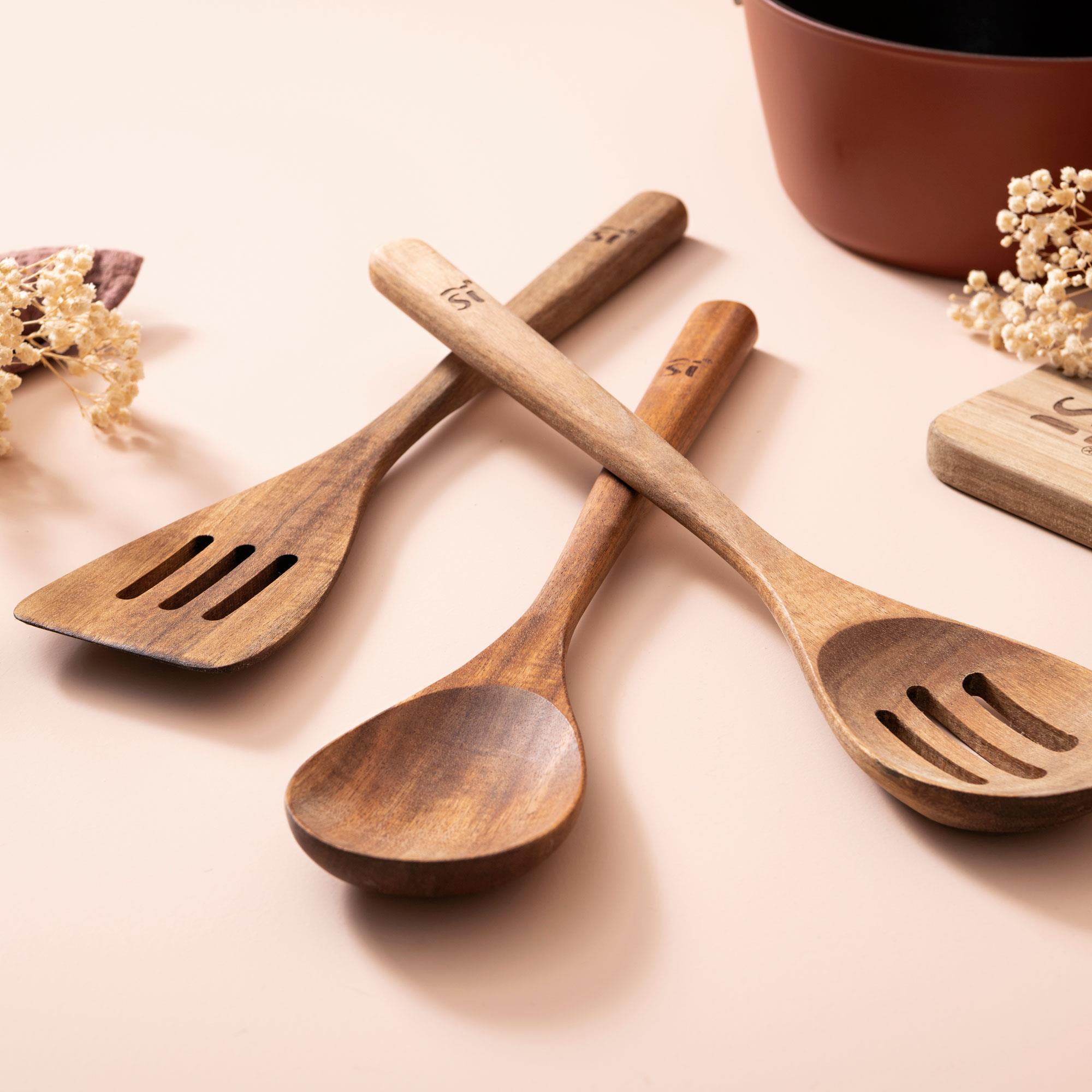 Utensilios de cocina de madera: espátula, cuchara y cuchara ranurada sobre una superficie rosa claro. Olla marrón y flores al fondo.