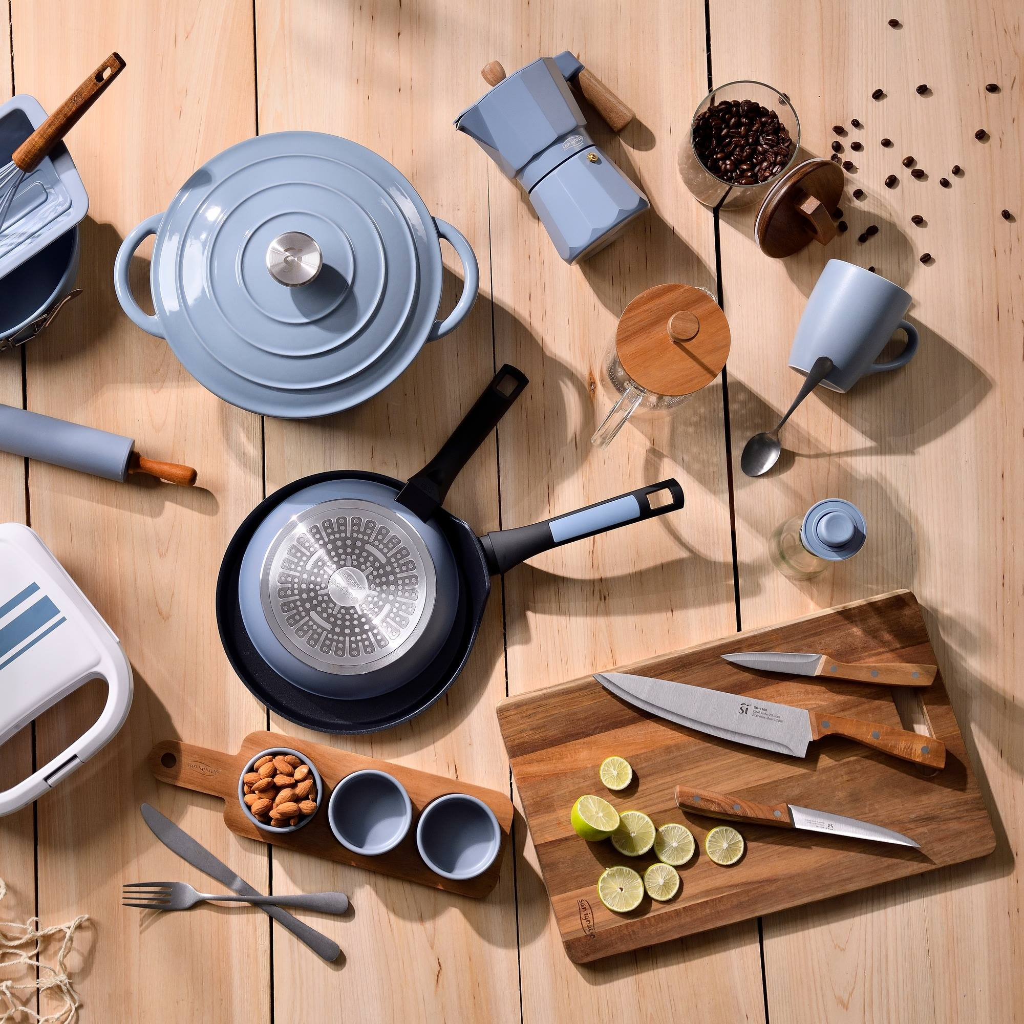 Vista aérea de artículos de cocina sobre una superficie de madera. Incluye ollas, sartenes, cafetera, cuchillos y alimentos.