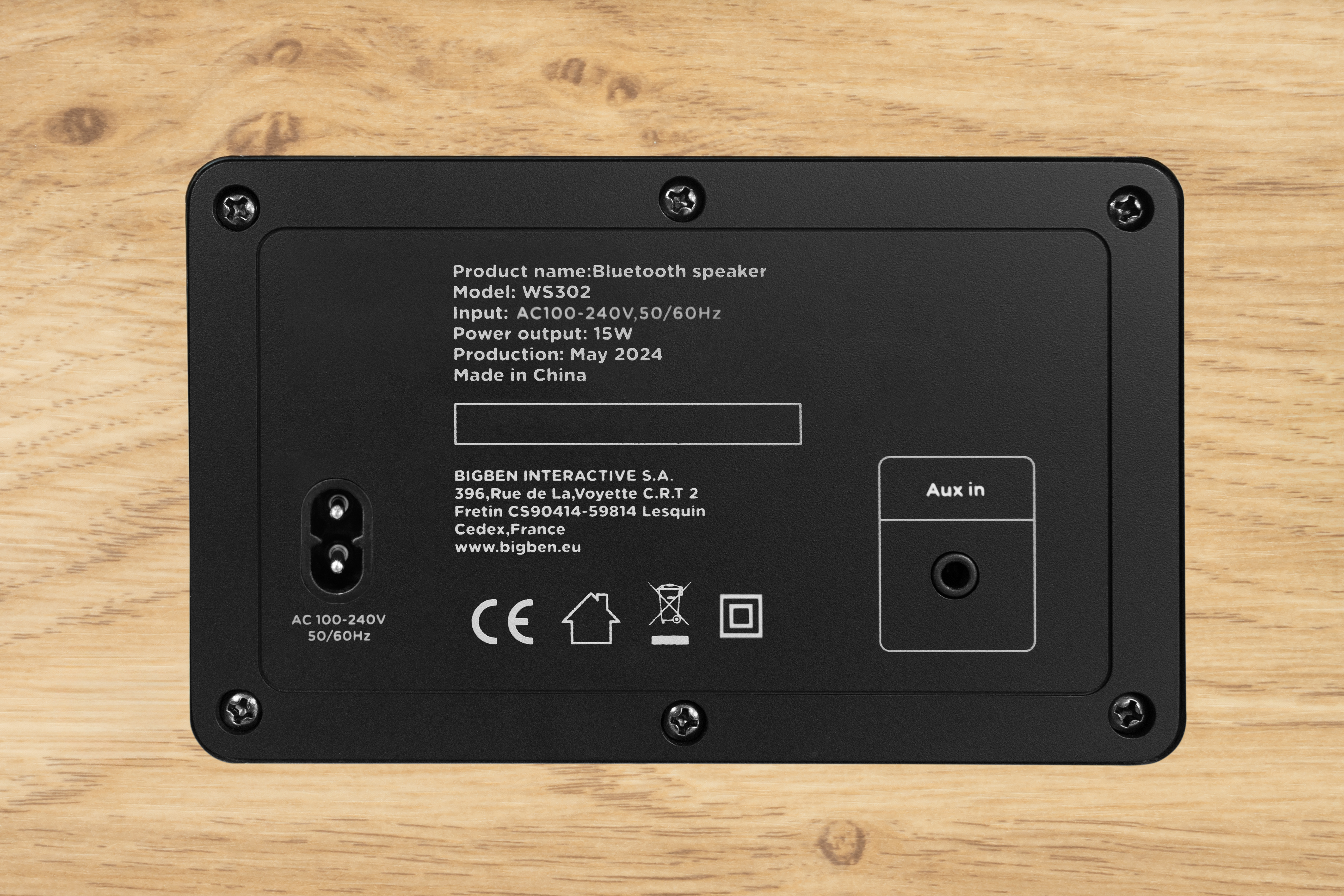 Czarny tył głośnika Bluetooth z tekstem na drewnianym tle. Zawiera wejście zasilania, Aux in i dane firmy.