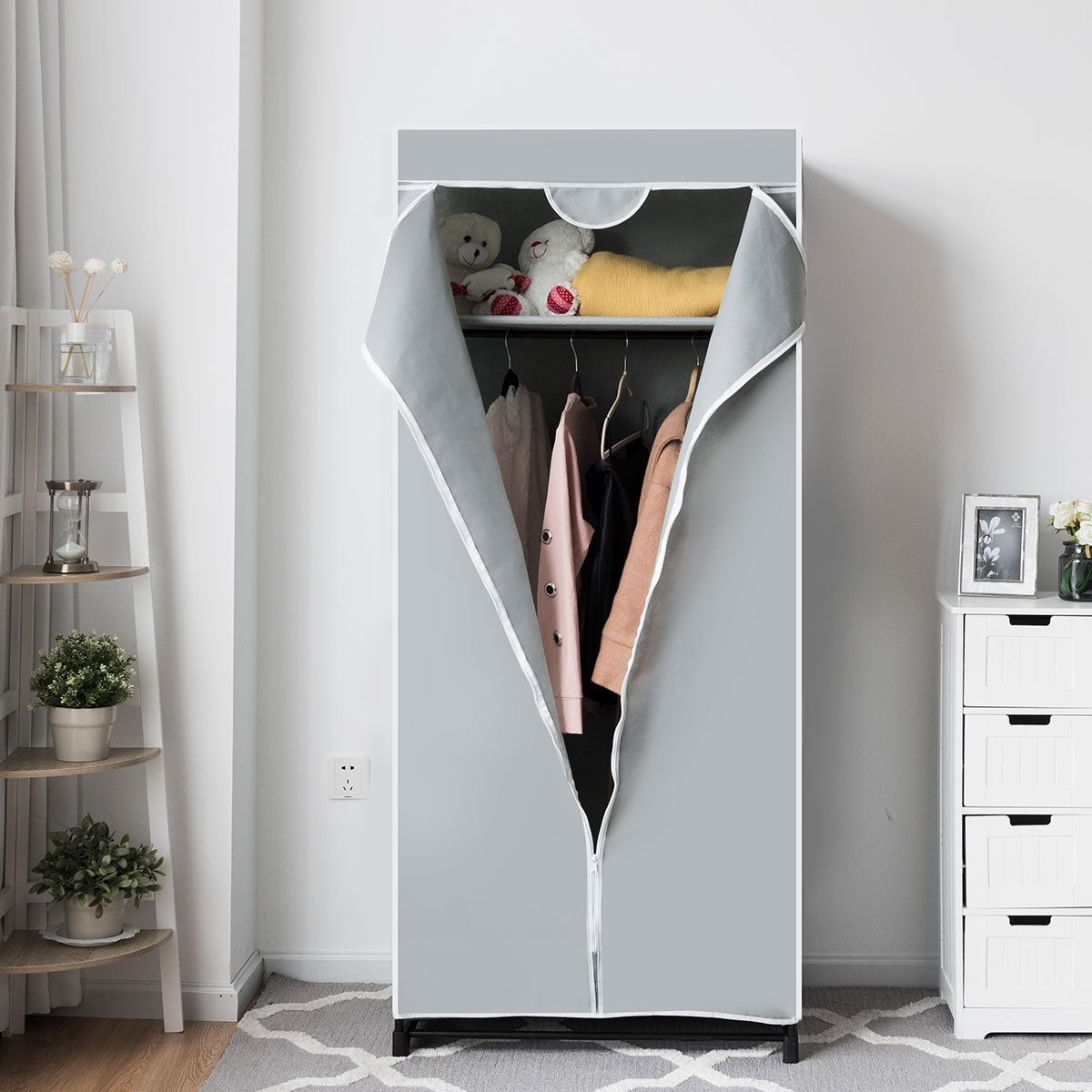 Un armario portátil gris con ropa colgada dentro. Un estante contiene peluches y una manta amarilla. Muebles y decoración blancos son visibles.