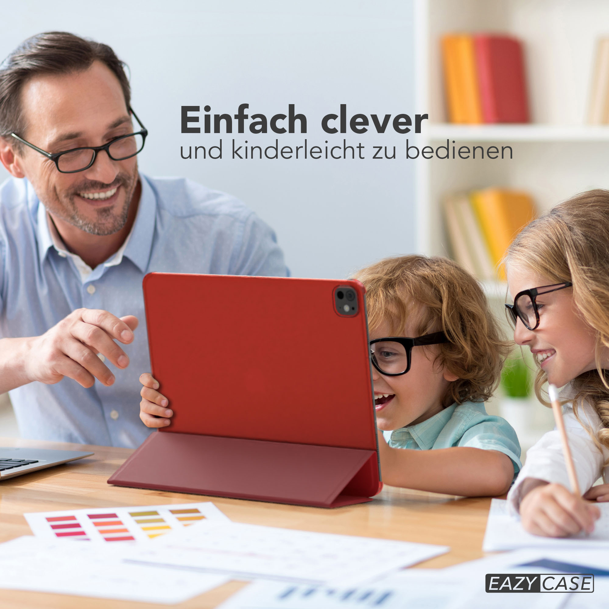 Ein Mann und zwei Kinder schauen auf ein rotes Tablet. Die Kinder tragen Brillen und lächeln. Ein Holztisch.