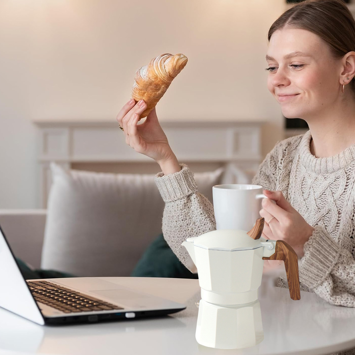 Eine Frau hält ein Croissant, eine Kaffeekanne und eine Tasse. Sie sitzt in der Nähe eines Laptops.
