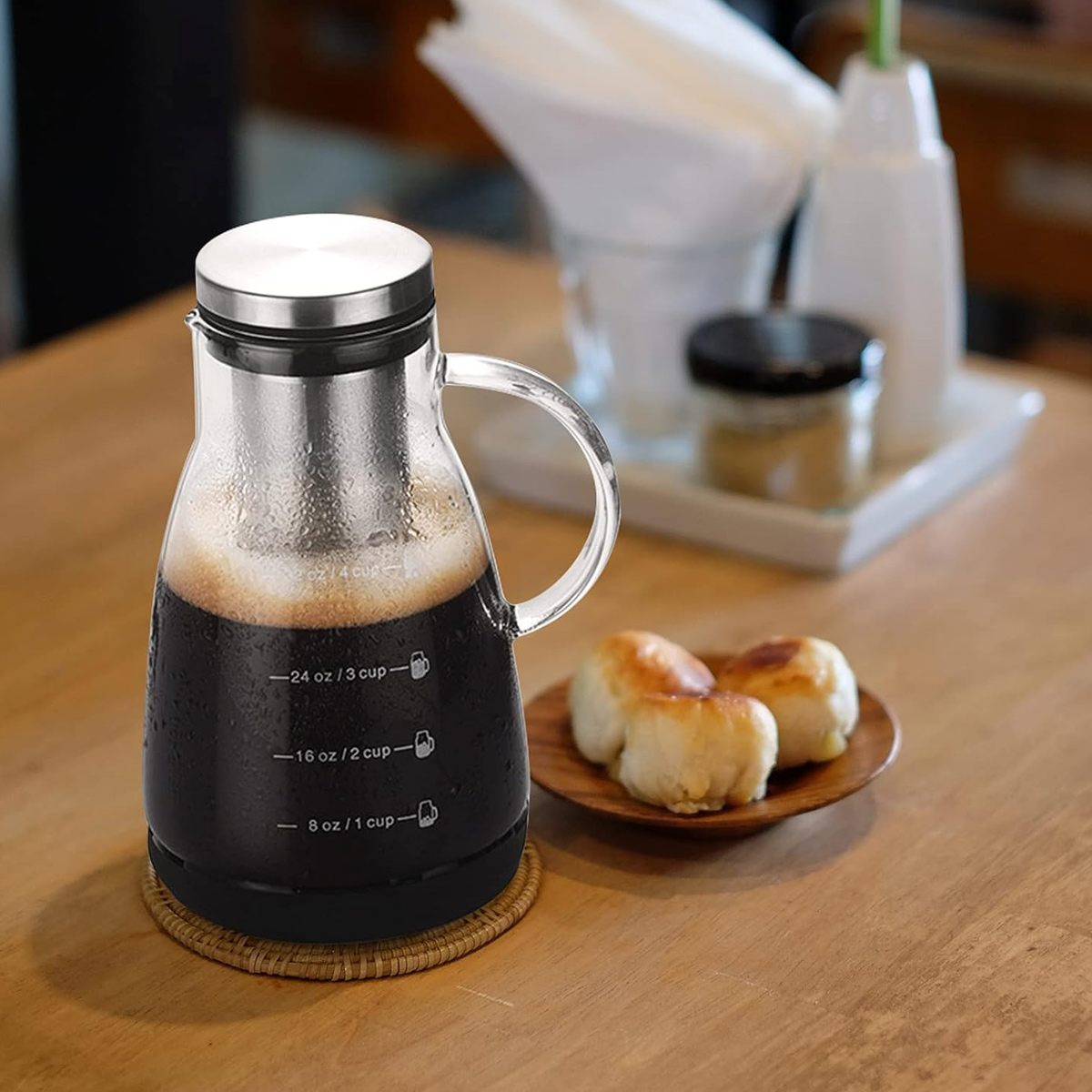 Eine Glaskaffeekaraffe mit Kaffee steht auf einem Holztisch. Auf einem Holzbrettchen liegen Brötchen, andere Gegenstände sind im Hintergrund.