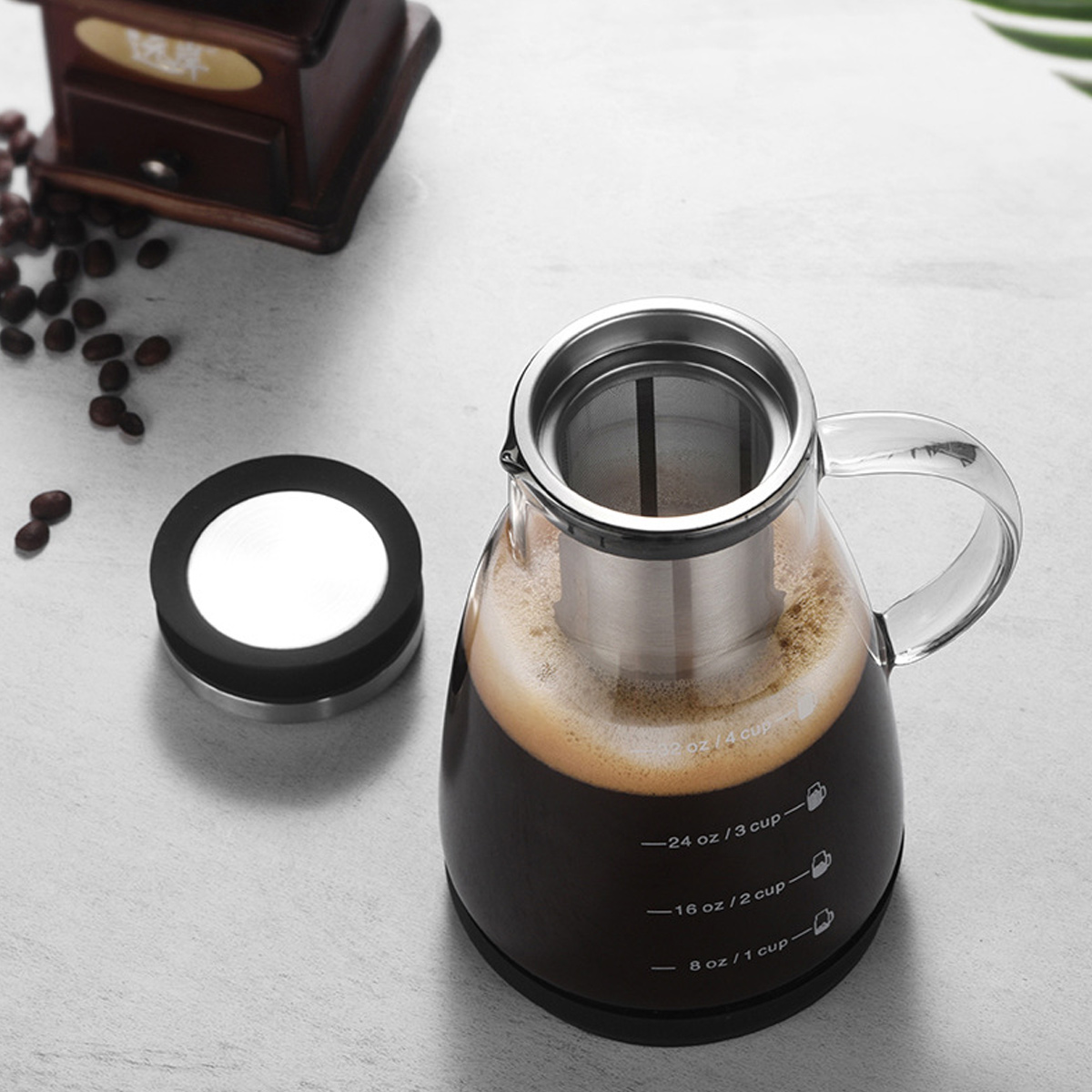 Ein Glas-Kaffeebereiter mit Kaffee, Deckel und einer Kaffeemühle im Hintergrund. Kaffeebohnen sind verstreut.