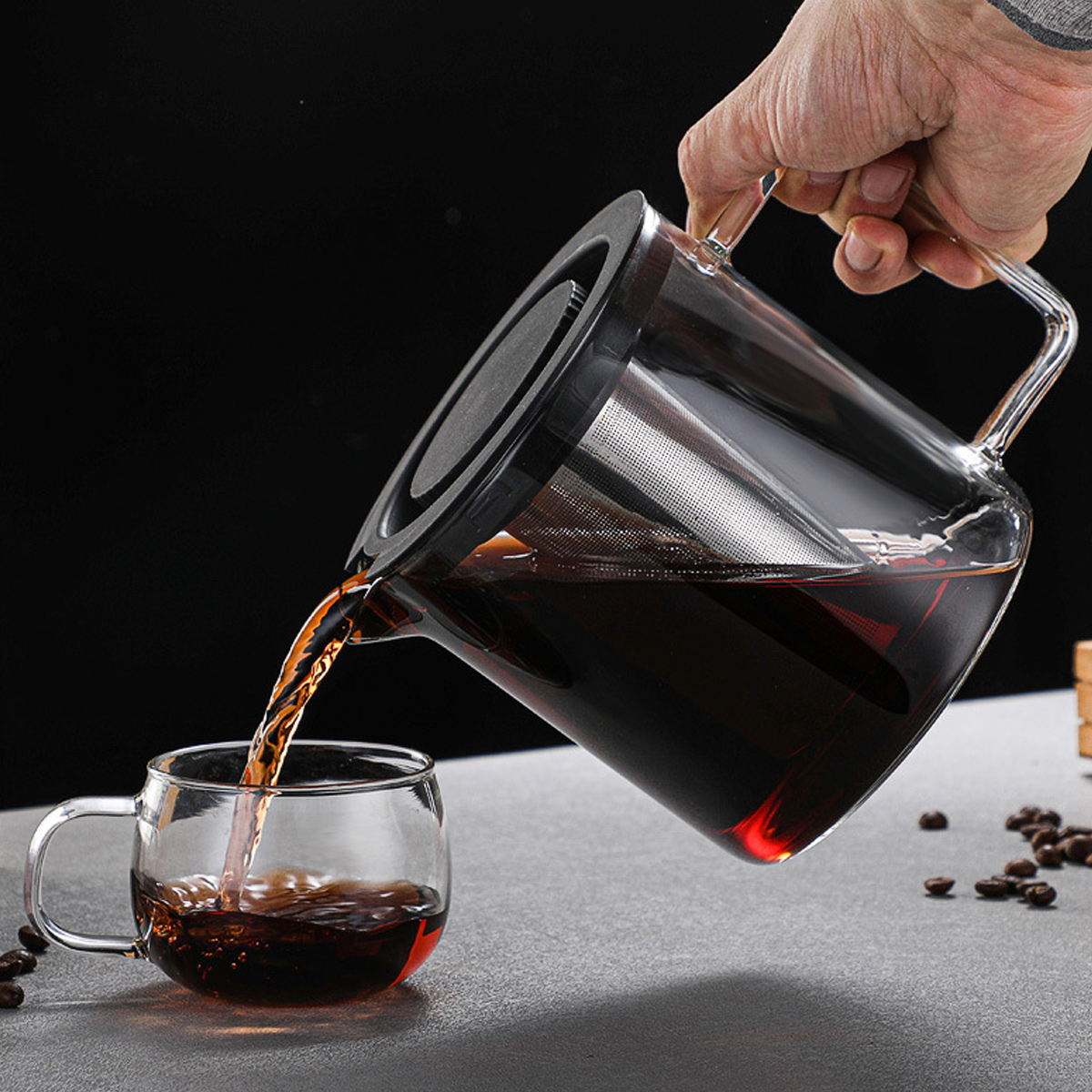 Eine Hand gießt Kaffee aus einer Glaskaraffe in eine Glastasse. Der Kaffee ist dunkel, und Kaffeebohnen sind auf dem Tisch verstreut.