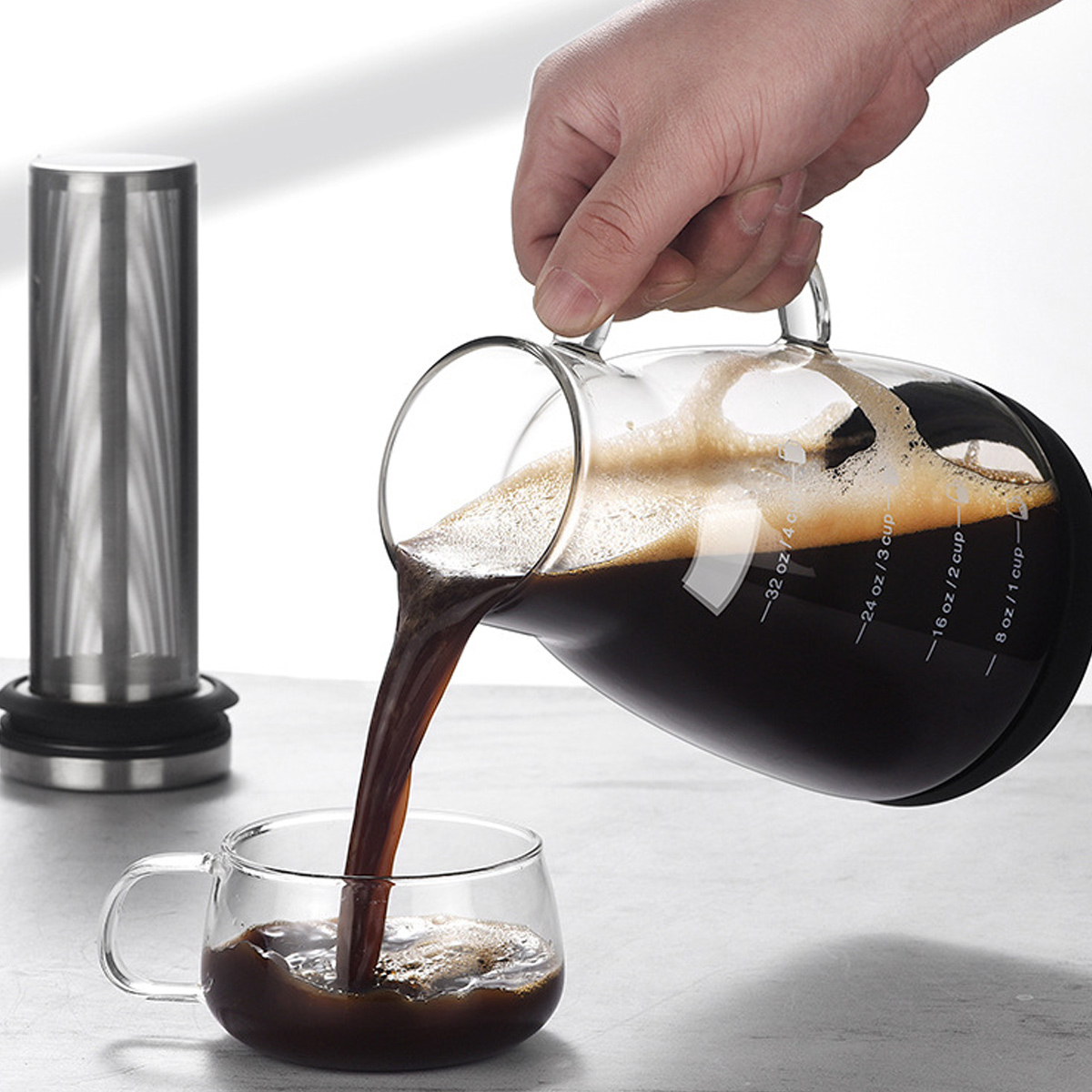 Eine Hand gießt Kaffee aus einer Glaskaraffe in eine Glastasse. Links befindet sich ein Edelstahlfilter.