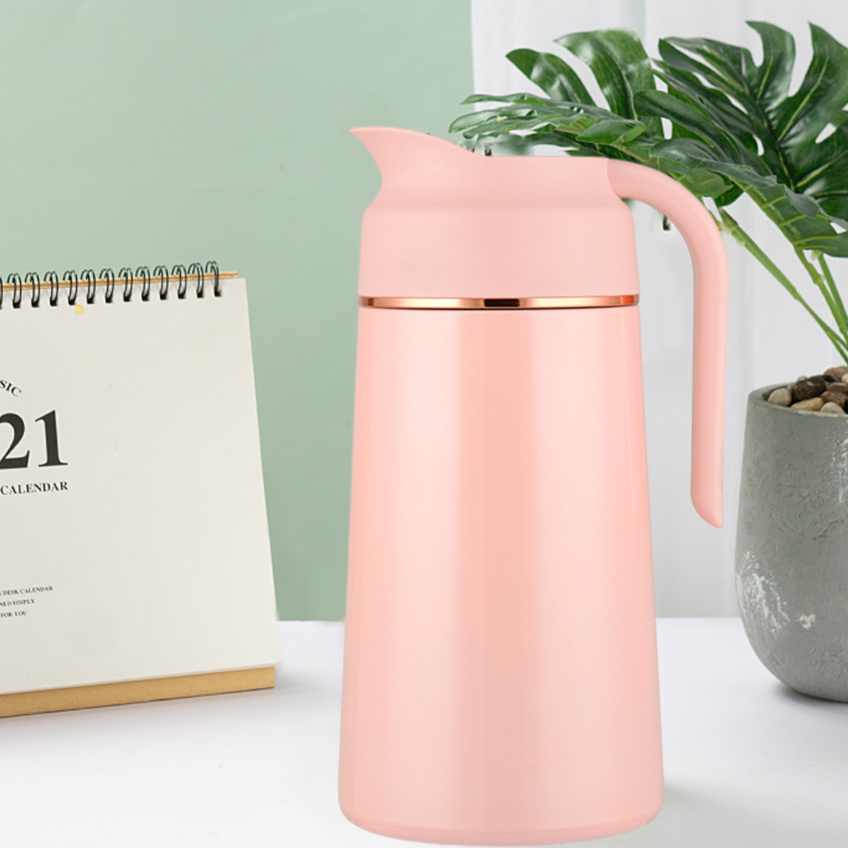 Eine rosa Thermoskanne mit Griff steht auf einer weißen Oberfläche. Ein Kalender und eine Topfpflanze sind im Hintergrund.