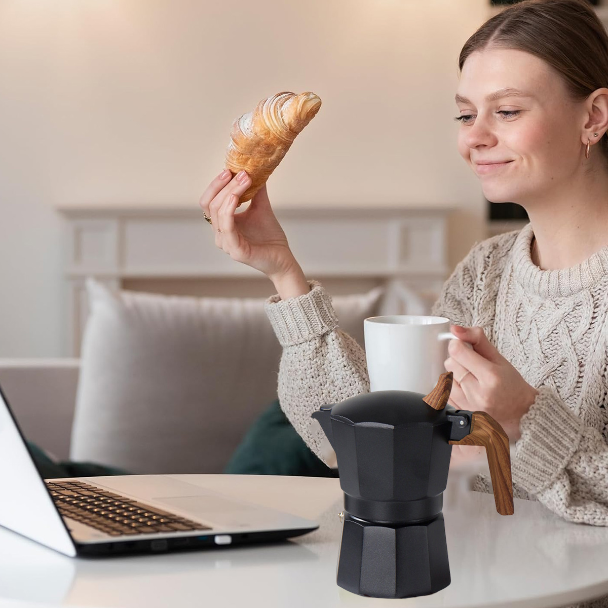 Frau hält Croissant, Kaffeetasse, Laptop und Kaffeemaschine auf einem Tisch. Im Hintergrund sind Sofa und Kamin.