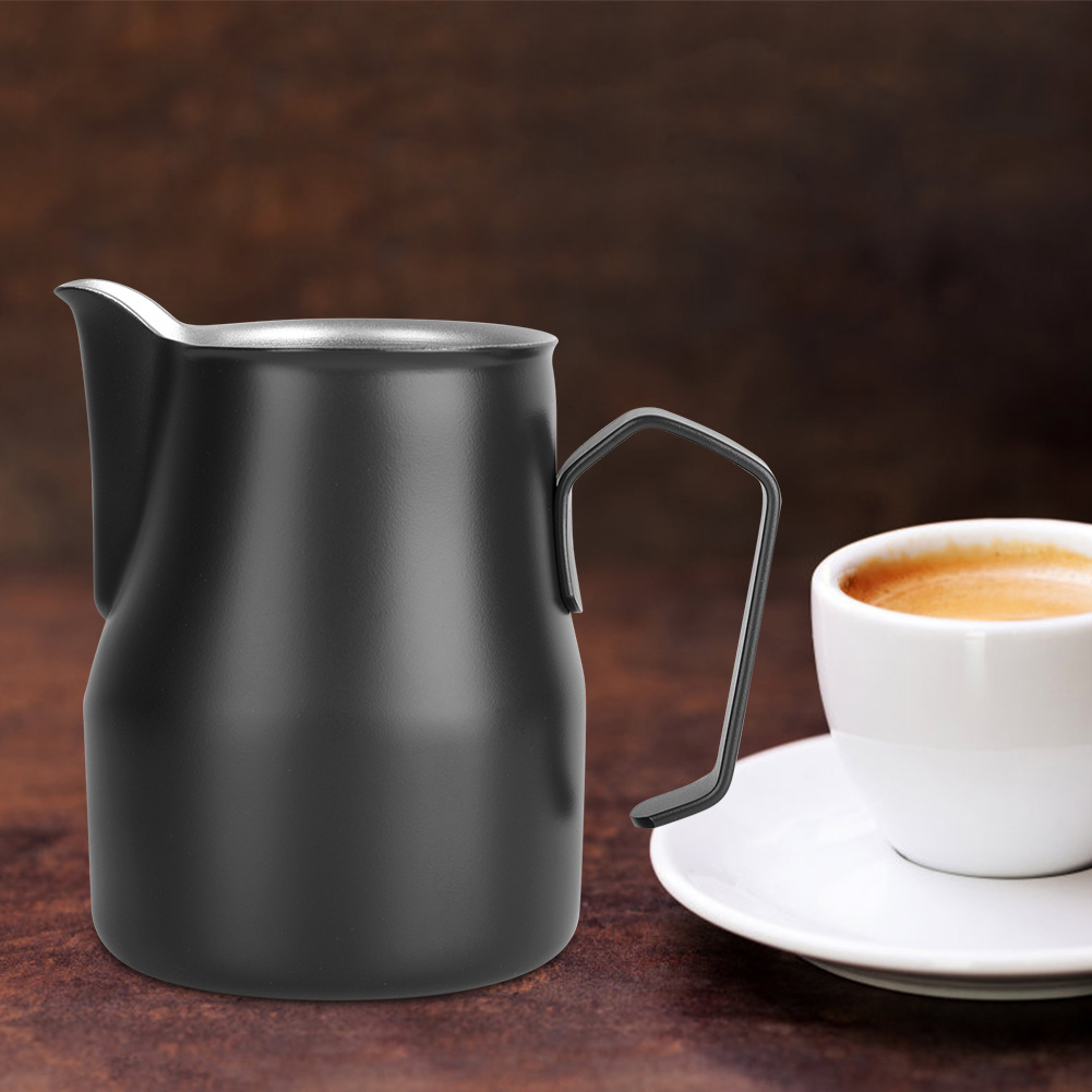 Schwarzer Milchaufschäumkrug und eine weiße Kaffeetasse mit Untertasse auf Holzoberfläche. Der Kaffee hat Crema.