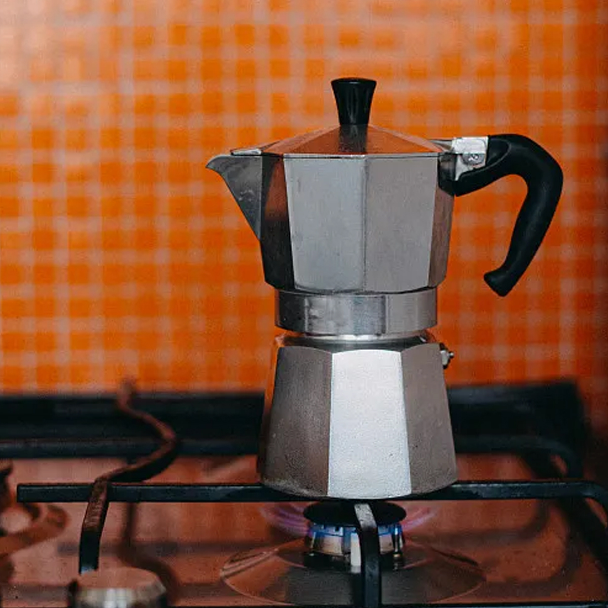 Ein silberner Espressokocher steht auf einem Gasbrenner. Der Hintergrund ist orangefarben kariert.