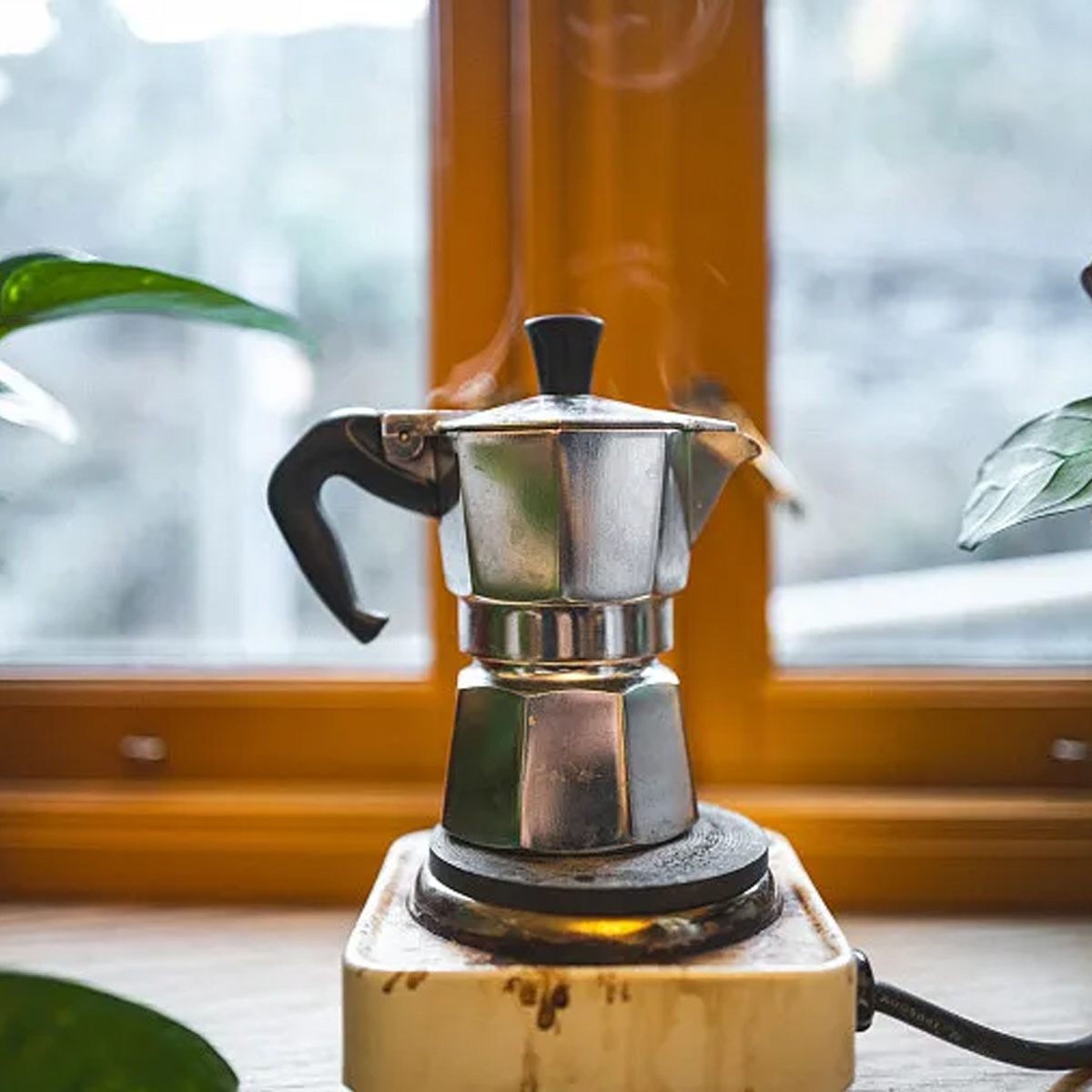 Ein Espressokocher auf dem Herd dampft. Er steht auf einem weißen Sockel in der Nähe eines Fensters. Pflanzen umrahmen das Fenster.