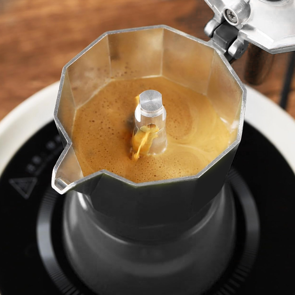 Eine Kaffeemaschine brüht Kaffee. Der Topf ist grau, der Kaffee braun. Er steht auf einer schwarz-weißen Oberfläche mit Holzgrund.