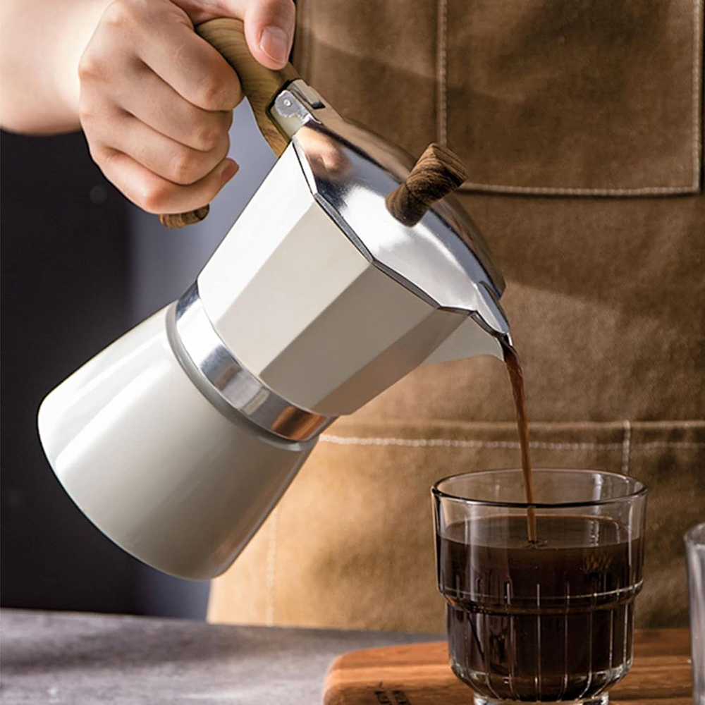 Eine Person gießt Kaffee aus einer weißen Mokkakanne in ein Glas. Kaffee ist dunkelbraun, die Kanne hat einen Holzgriff.