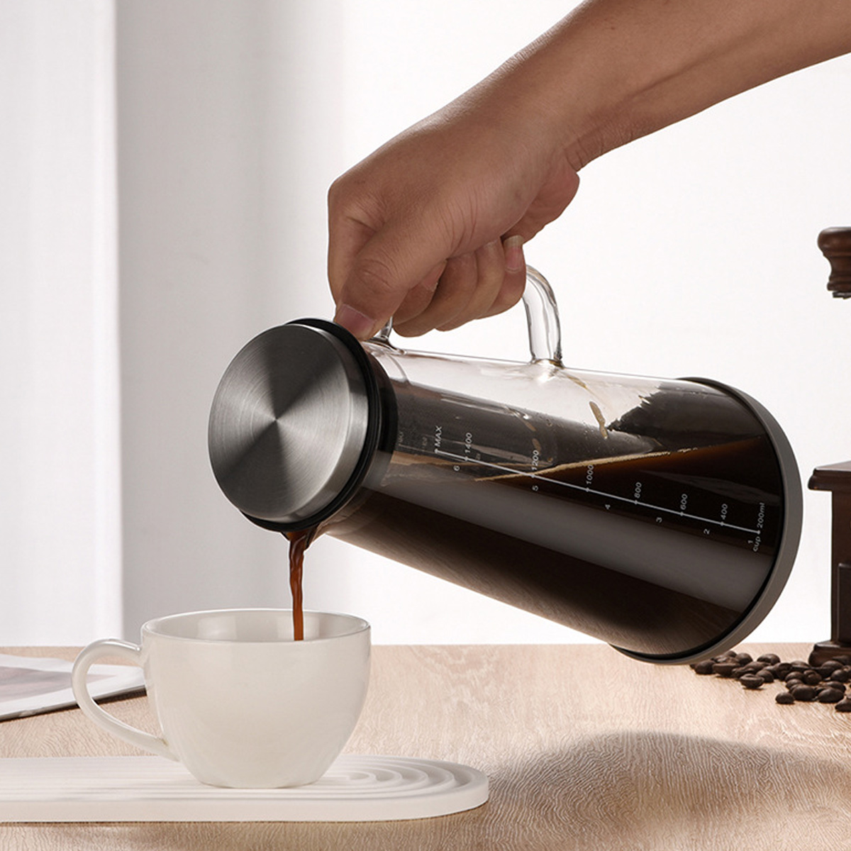 Eine Hand gießt Kaffee aus einer Glaskaraffe in eine weiße Tasse. Kaffeebohnen und eine Mühle sind im Hintergrund zu sehen.