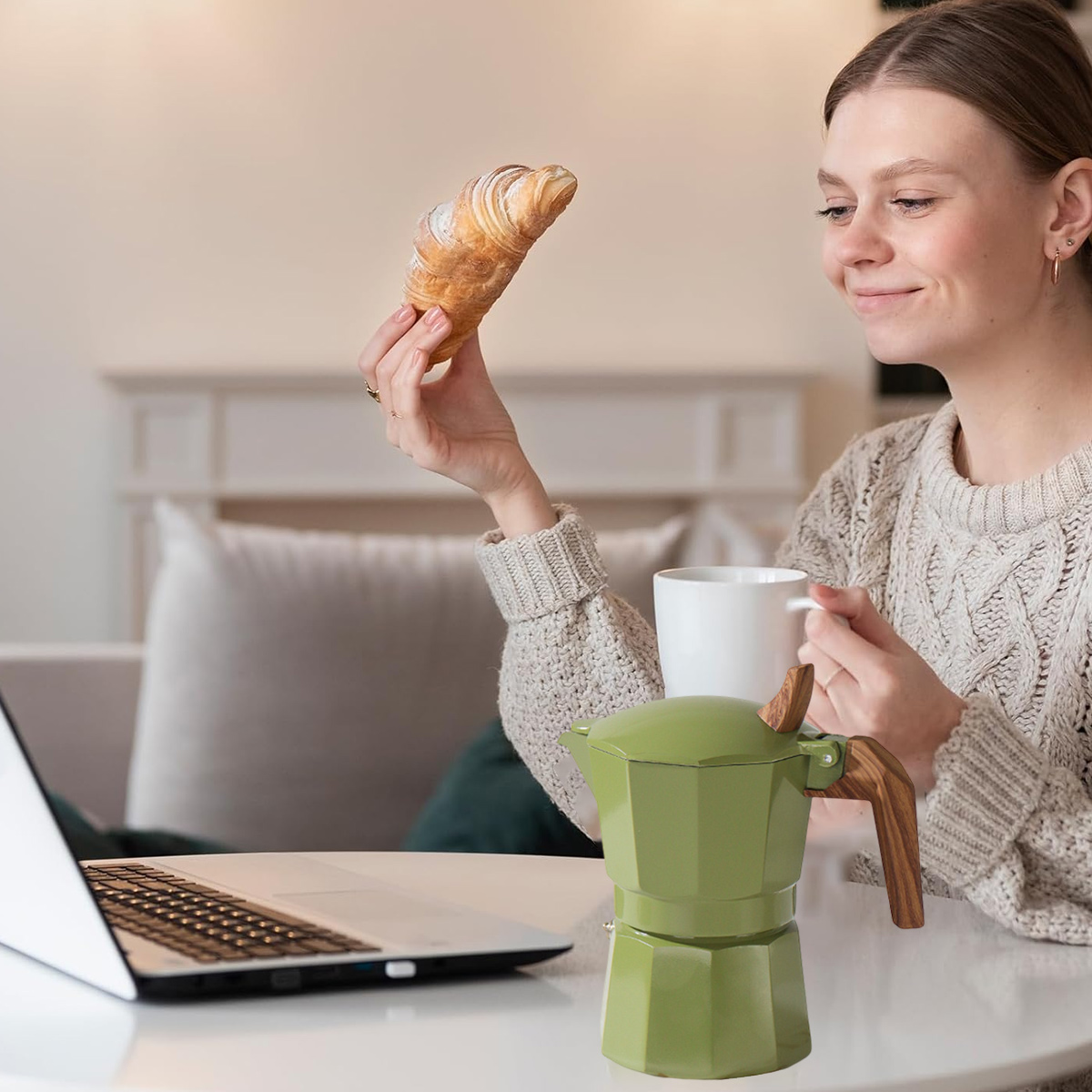 Frau hält Croissant, Kaffee und grüne Kaffeemaschine. Laptop auf dem Tisch. Hintergrund ist ein Wohnzimmer mit Sofa.
