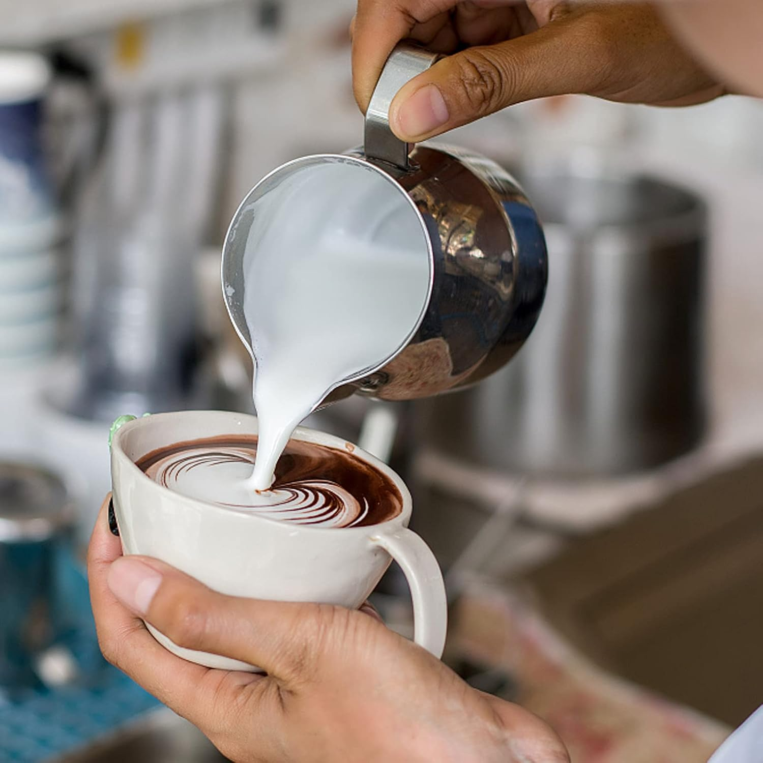 Hände gießen Milch aus einem Metallkrug in eine Tasse Kaffee und erzeugen ein Design. Die Tasse wird in einer anderen Hand gehalten.