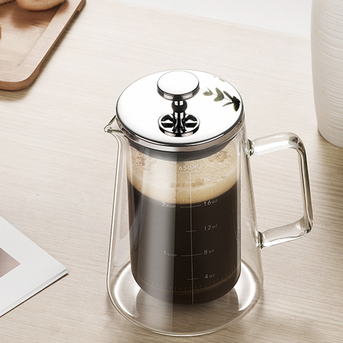 Eine klare Glaskaffeepresse, gefüllt mit dunklem Kaffee. Sie hat einen silbernen Deckel und einen Griff. Sie steht auf einer hellen Holzoberfläche.