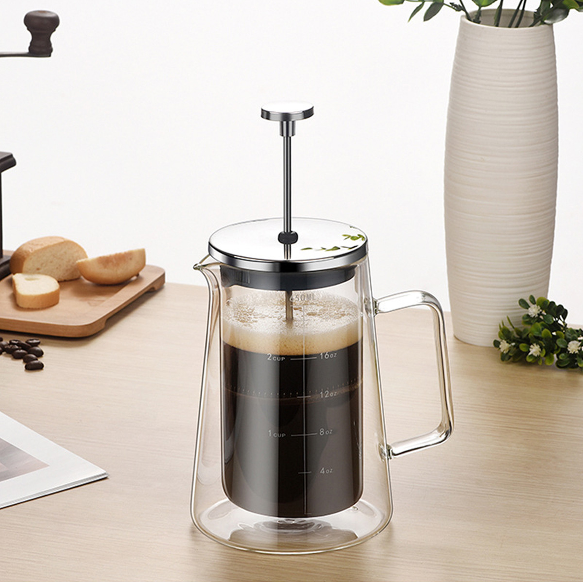 Eine French Press mit Kaffee steht auf einem Holztisch. Brot und Kaffeebohnen sind in der Nähe. Eine Vase mit einer Pflanze steht hinten.