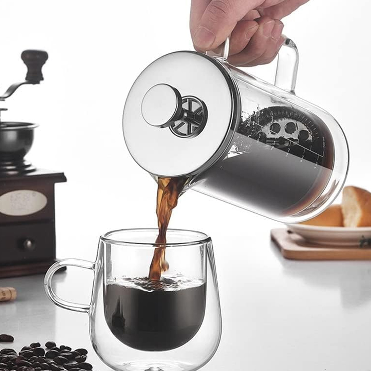 Kaffee wird aus einer Glas-French-Press in eine Glastasse gegossen. Im Hintergrund befindet sich eine Kaffeemühle mit Kaffeebohnen.