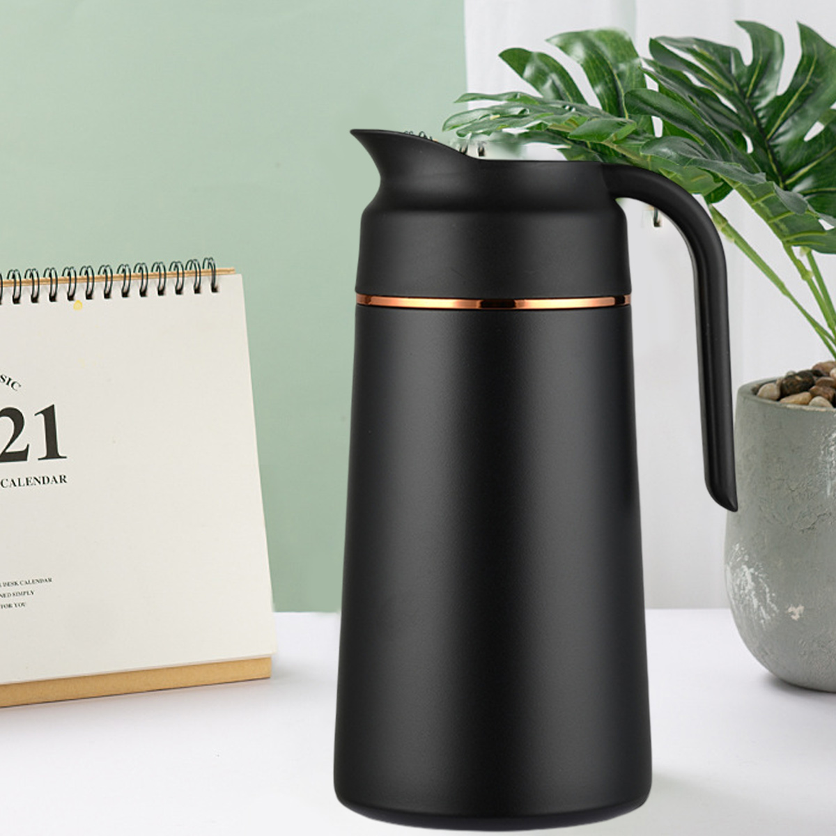 Eine schwarze Thermoskanne mit goldenem Band steht neben einem Kalender und einer Topfpflanze.