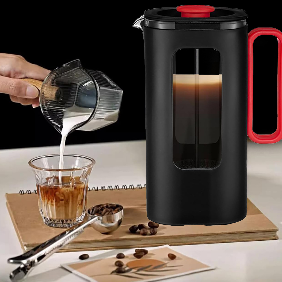 Milch wird in ein Glas Kaffee gegossen. Eine French Press und Kaffeebohnen liegen auf einem Notizbuch.