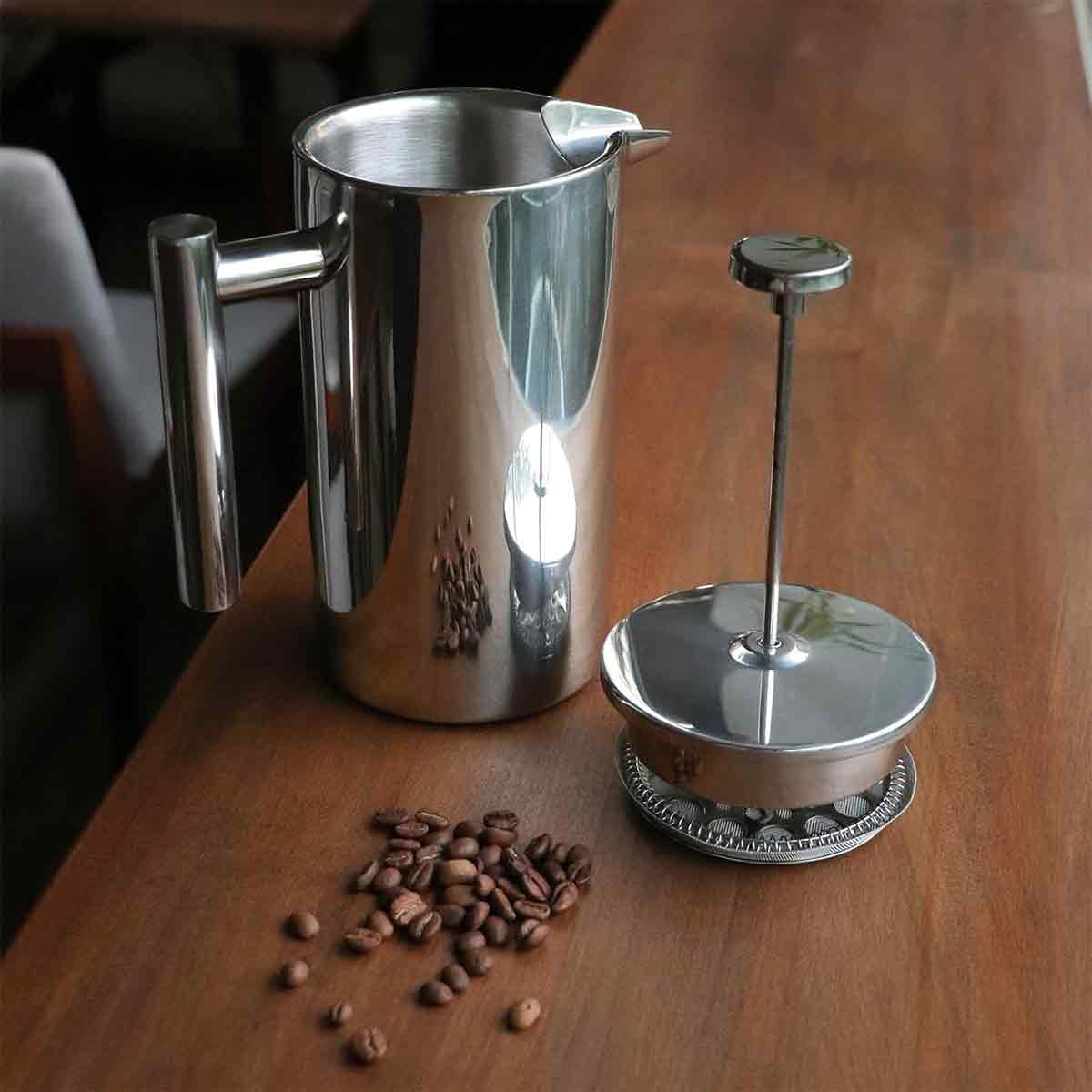 Eine auseinandergebaute French Press steht auf einem Holztisch. Kaffeebohnen sind in der Nähe verstreut. Die Presse ist silber und glänzend.