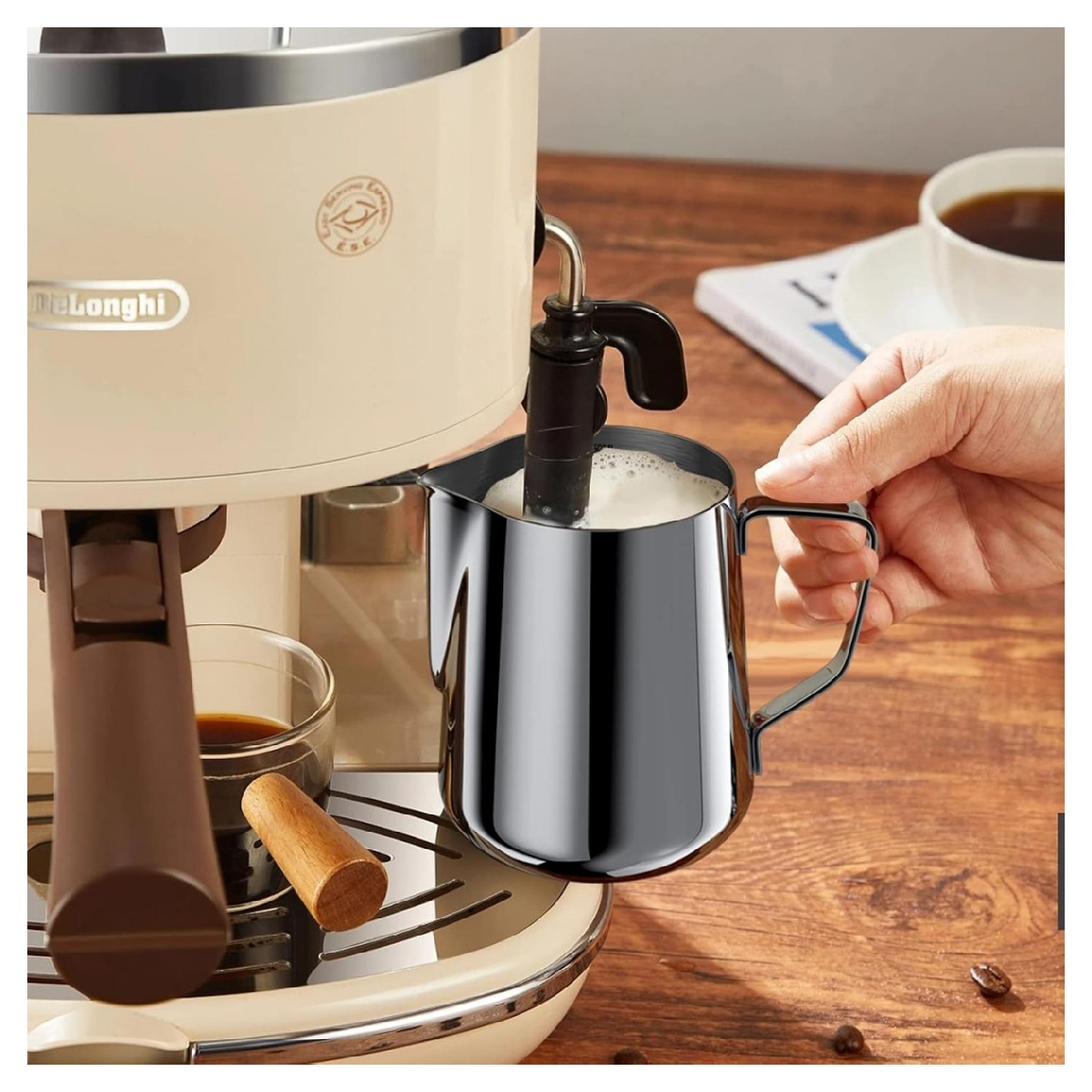 Eine Person schäumt Milch mit einer Dampflanze an einer Espressomaschine auf. Ein Krug aus Edelstahl enthält die Milch. Kaffee ist in einem Glas.