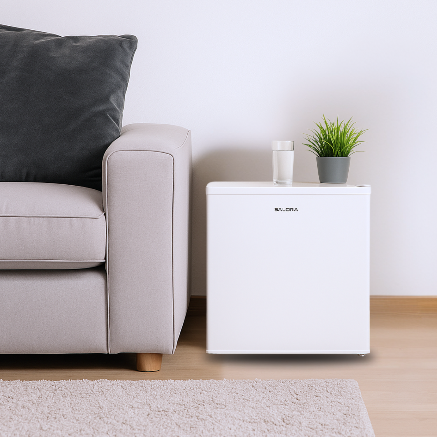 A white mini-fridge next to a sofa with a gray pillow. A glass of water and a plant sit on the fridge.