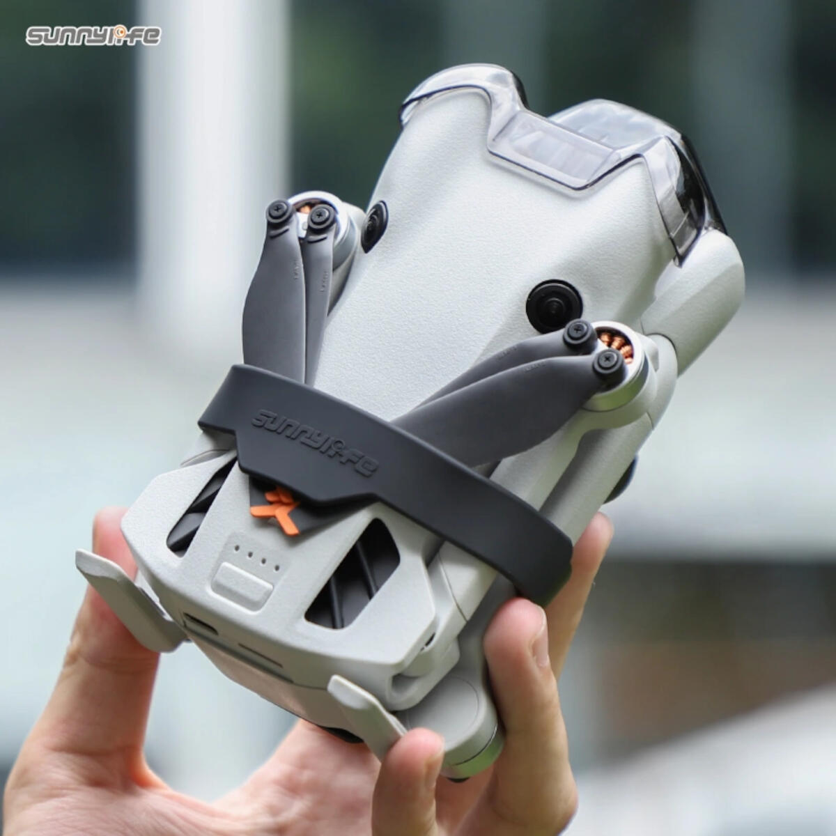 Un drone gris avec des hélices noires et un couvercle de protection est tenu par une main. Le drone est contre un fond vert flou.