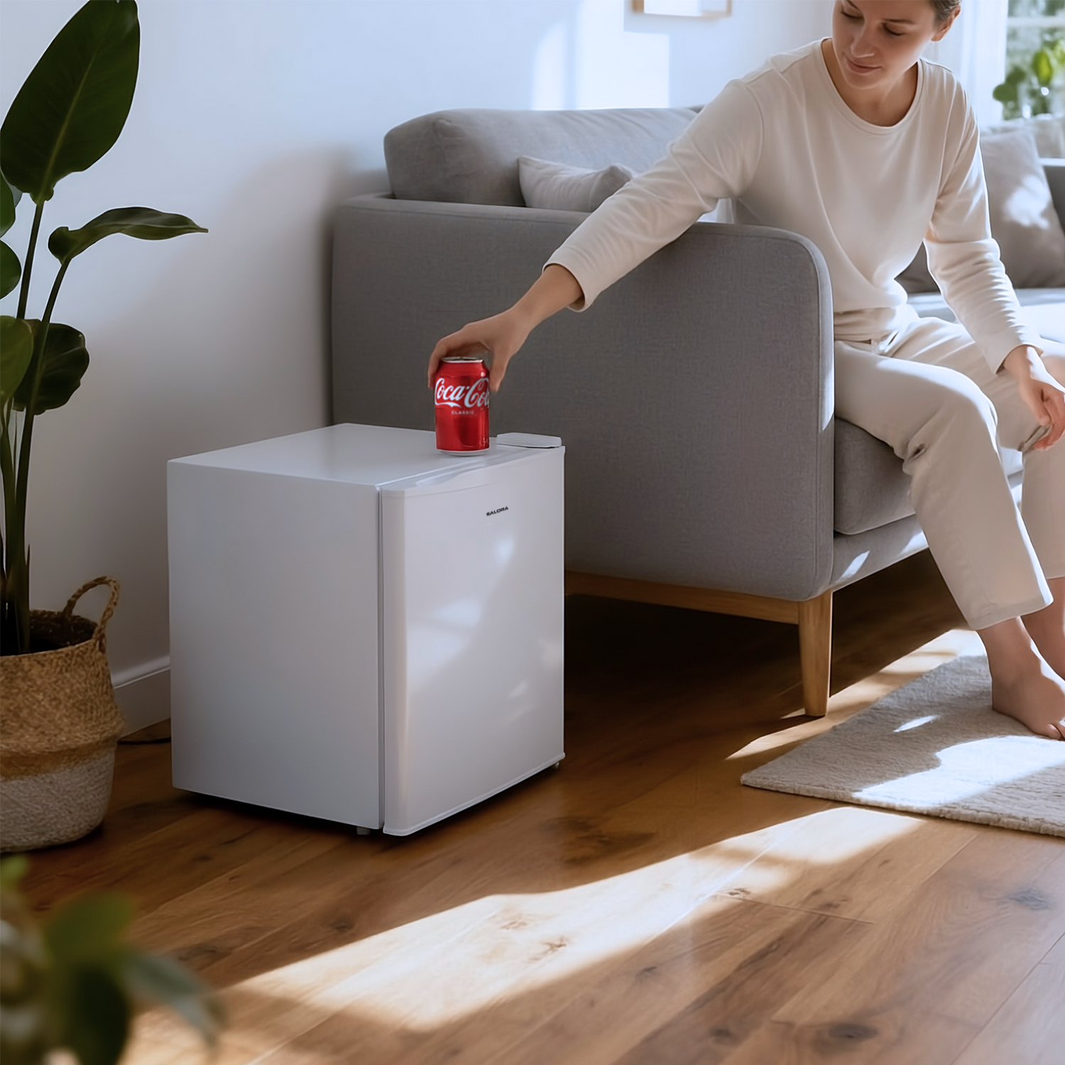 Woman reaching for a Coca-Cola can on a white mini-fridge. Beside a sofa and plant. Wooden floor.