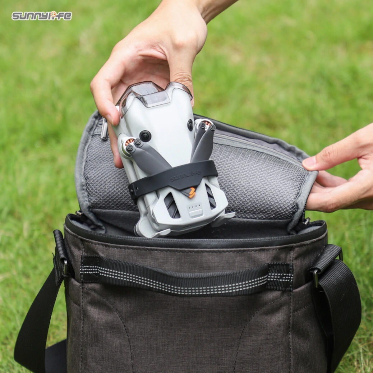 Une personne place un drone gris avec des hélices noires dans un sac gris foncé. Herbe verte en arrière-plan flou.