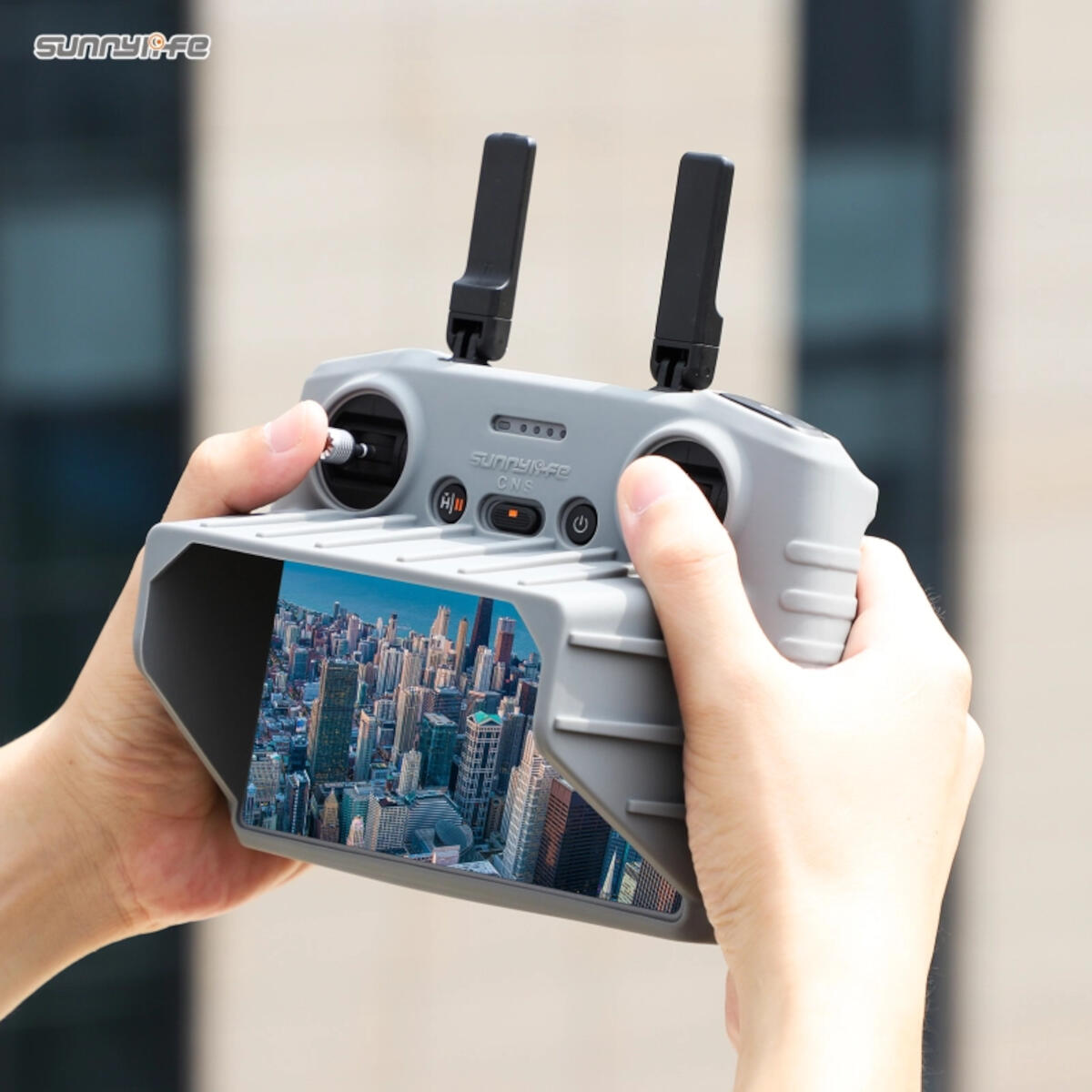 A person holds a drone controller, displaying a city view. The controller is gray, has antennas and buttons. The background is blurred.