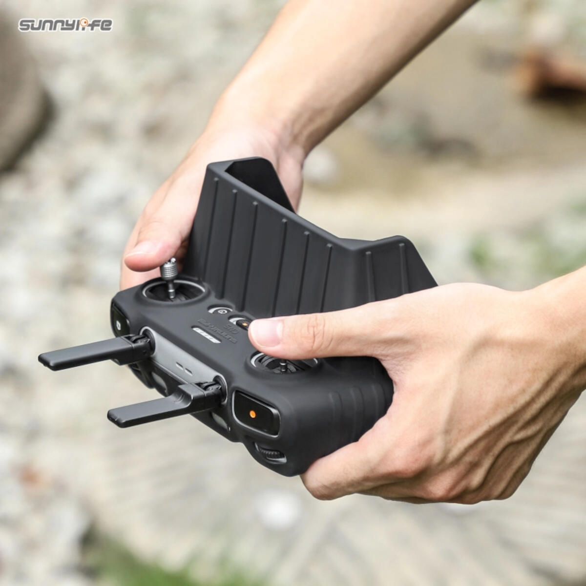 Hands holding a black drone controller with antennas extended. The controller is in a black protective case. Blurred green background.