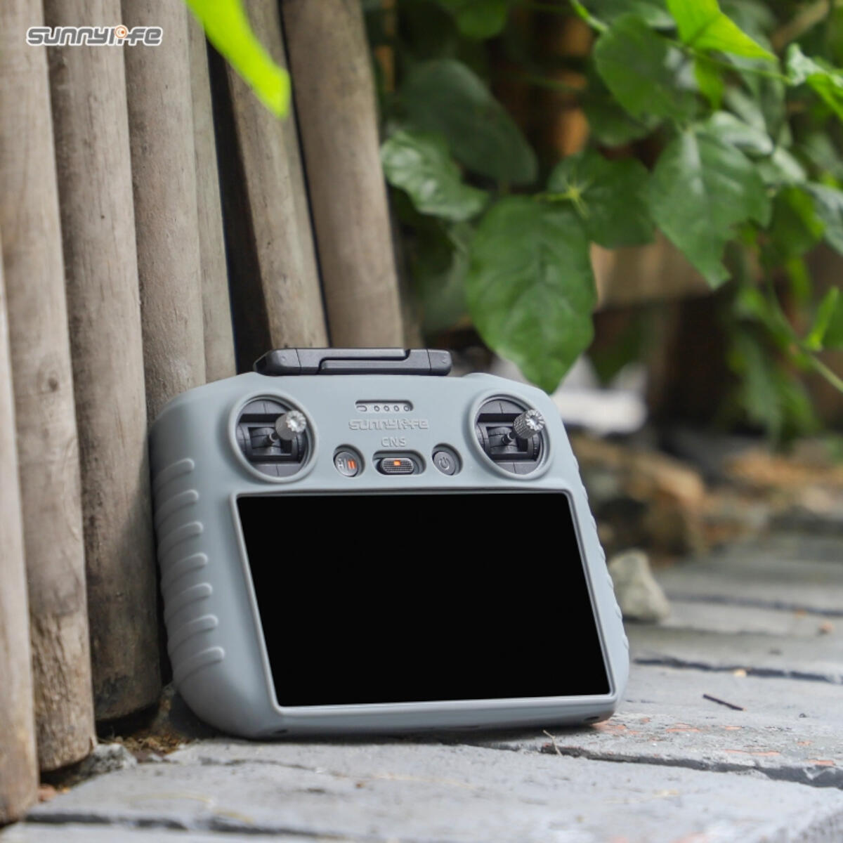 A gray drone controller is propped against wooden planks. Green leaves and a stone surface are in the background.