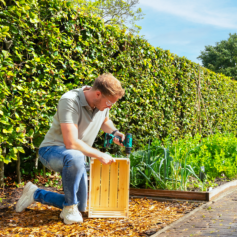 Man knielt, boort een houten kist in een tuin. Groene haag op de achtergrond, blauwe lucht. Houtsnippers en planten eromheen.