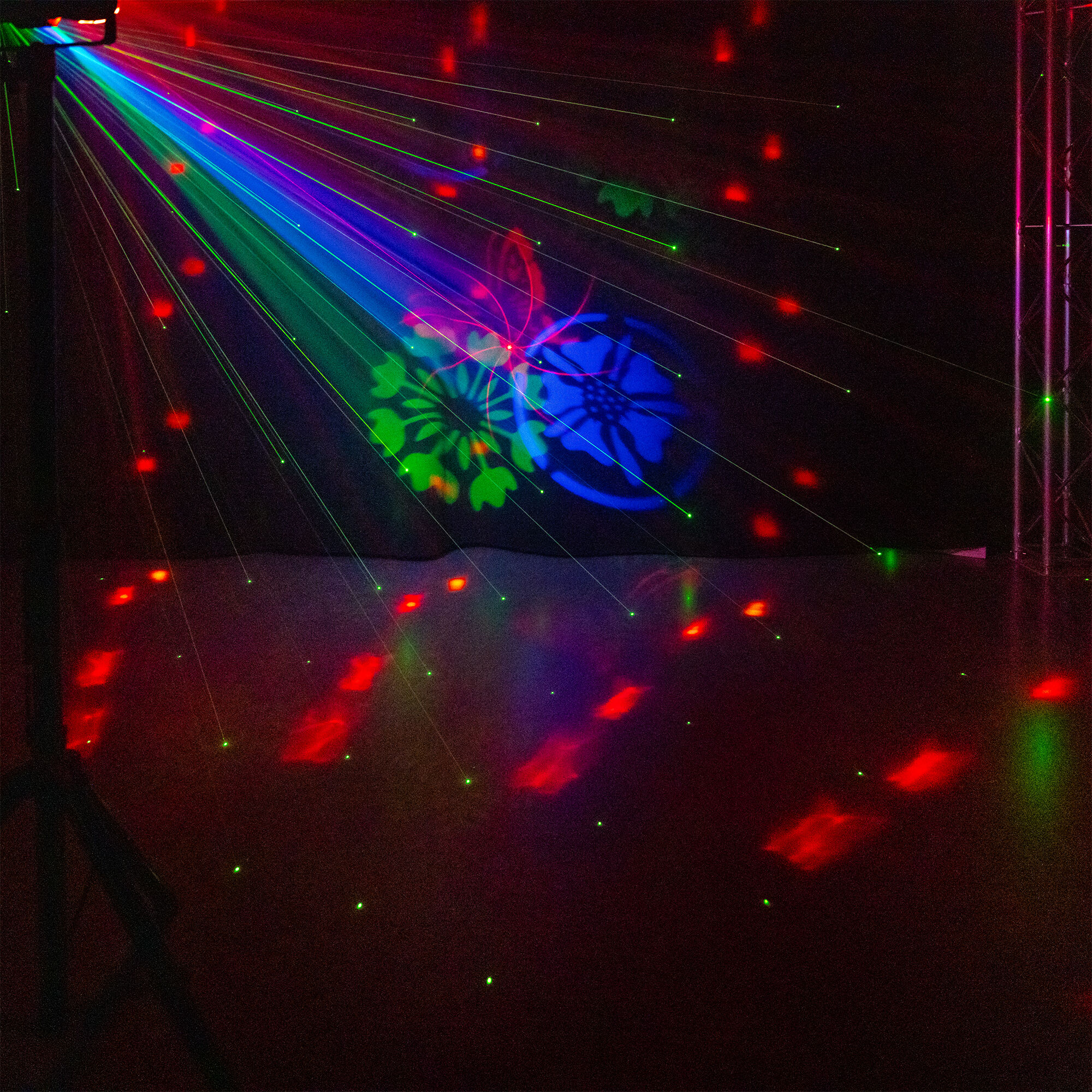 Un espectáculo láser ilumina una habitación oscura con luces verdes, azules y rojas. Patrones florales y haces crean una escena vibrante.