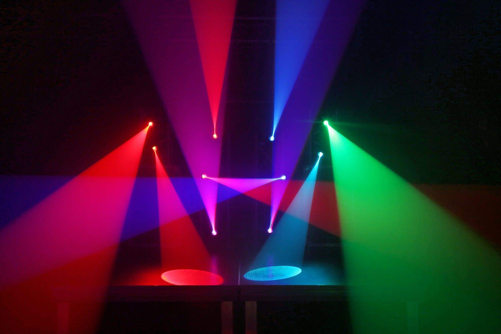 Luces de escenario coloridas iluminan un espacio oscuro. Rayos rojos, azules, verdes y morados convergen, creando una exhibición vibrante.