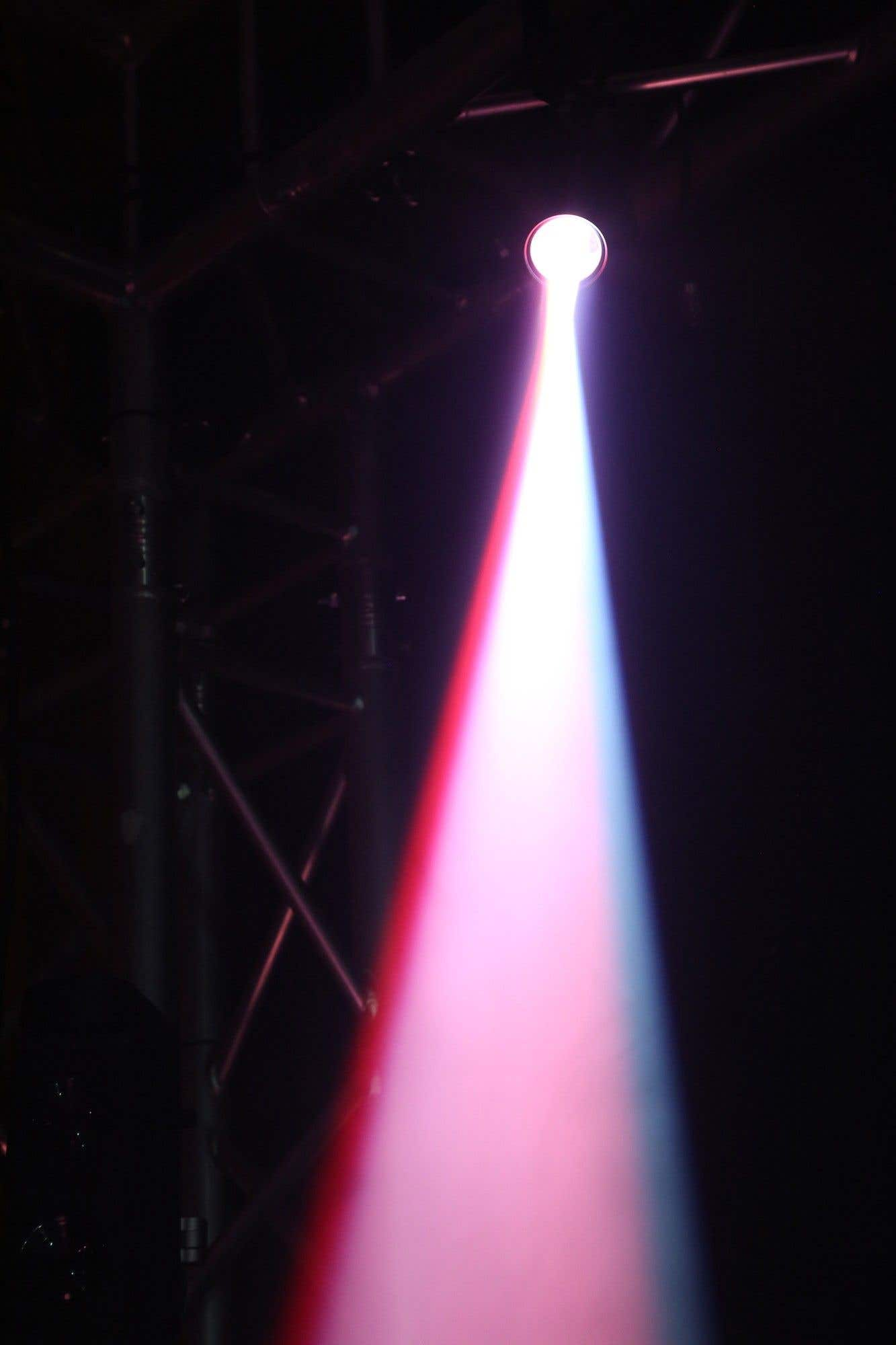 Un foco brilla hacia abajo, con tonos rojos y azules. El haz ilumina un espacio oscuro. Estructuras metálicas visibles a la izquierda.