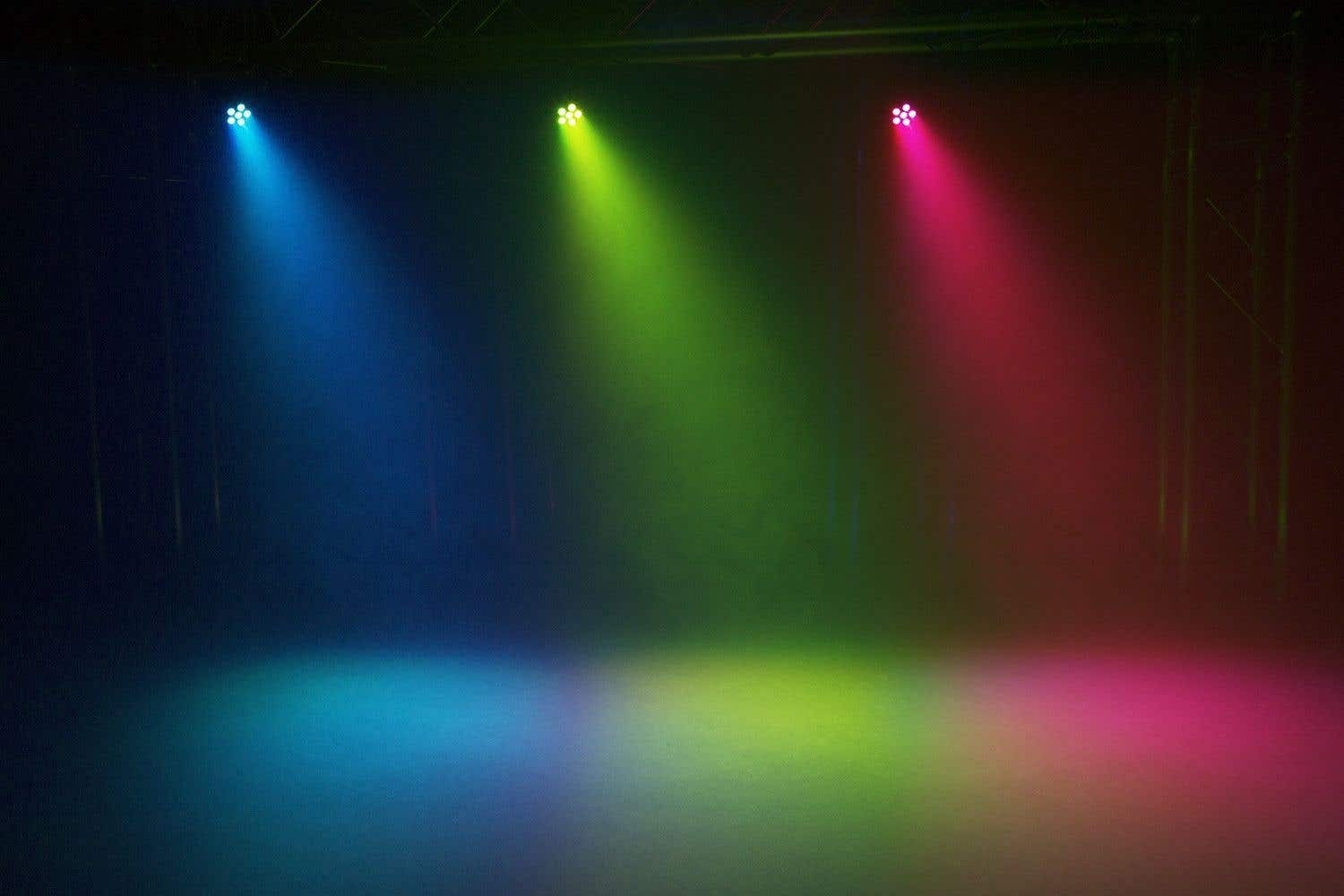 Tres luces de escenario proyectan haces azules, verdes y rosas sobre un suelo de escenario humeante, creando un efecto atmosférico colorido.