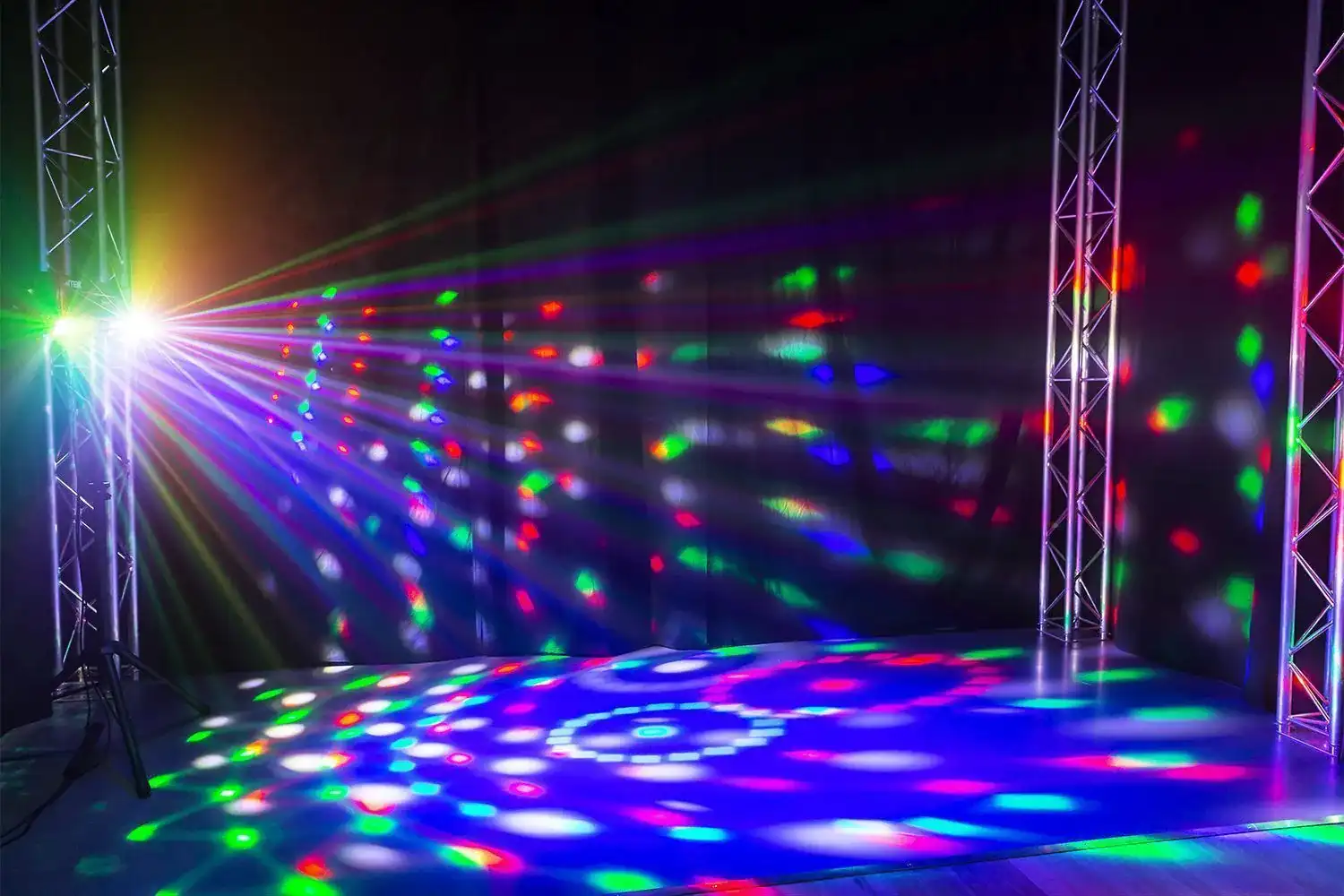 Un escenario iluminado con focos de colores. Estructuras metálicas enmarcan la escena. Luces proyectan patrones en el suelo.