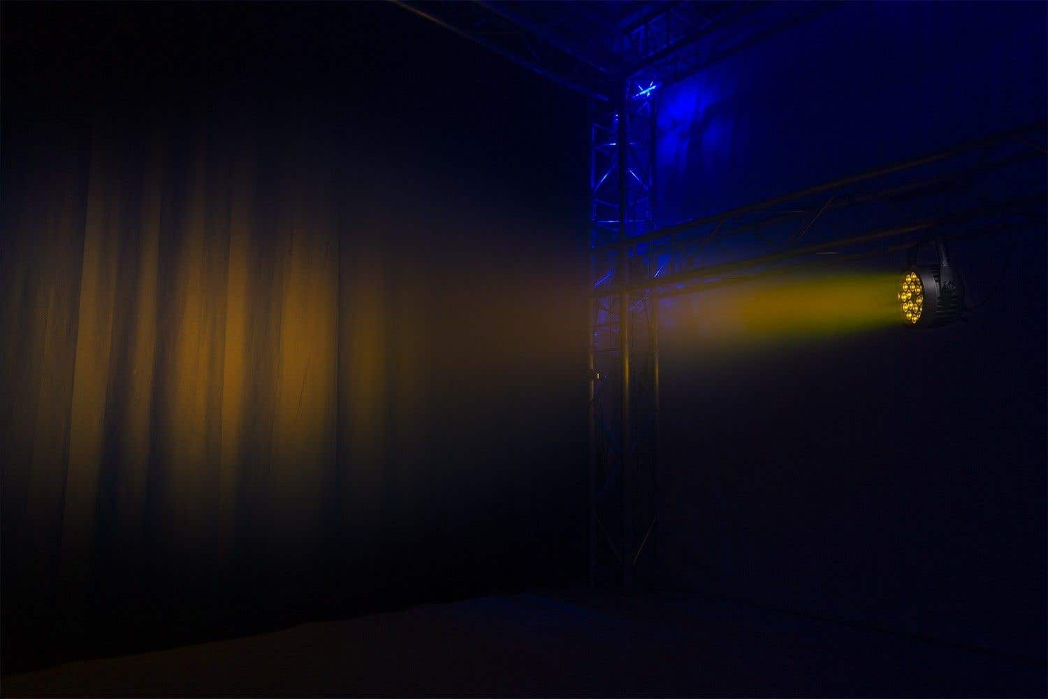 Un escenario con un foco que emite luz amarilla. Fondo oscuro con cortinas. Estructura metálica a la derecha.