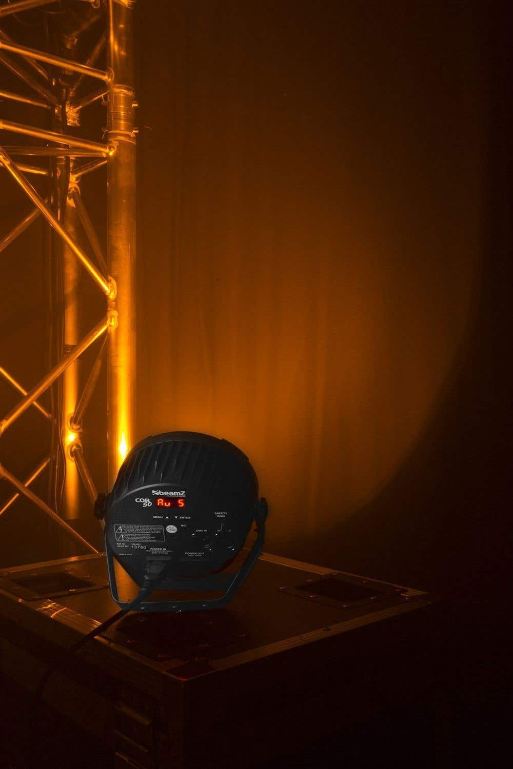 Una luz de escenario Beamz está sobre una superficie oscura. Una estructura metálica está en el fondo, iluminada con luz naranja.