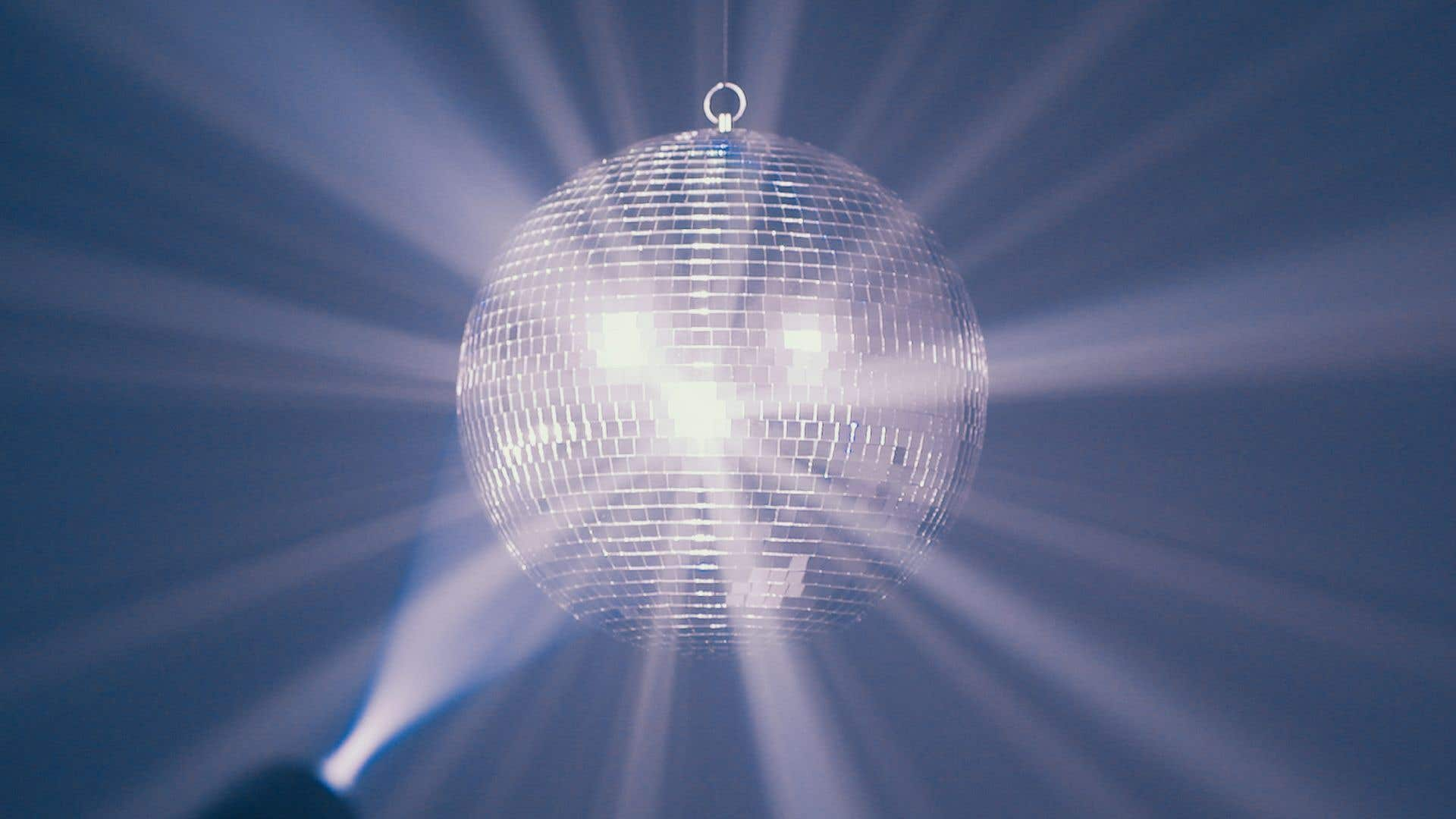 Una bola de discoteca cuelga, reflejando luz. Rayos blancos irradian hacia afuera. El fondo es un gradiente de tonos azules y morados.