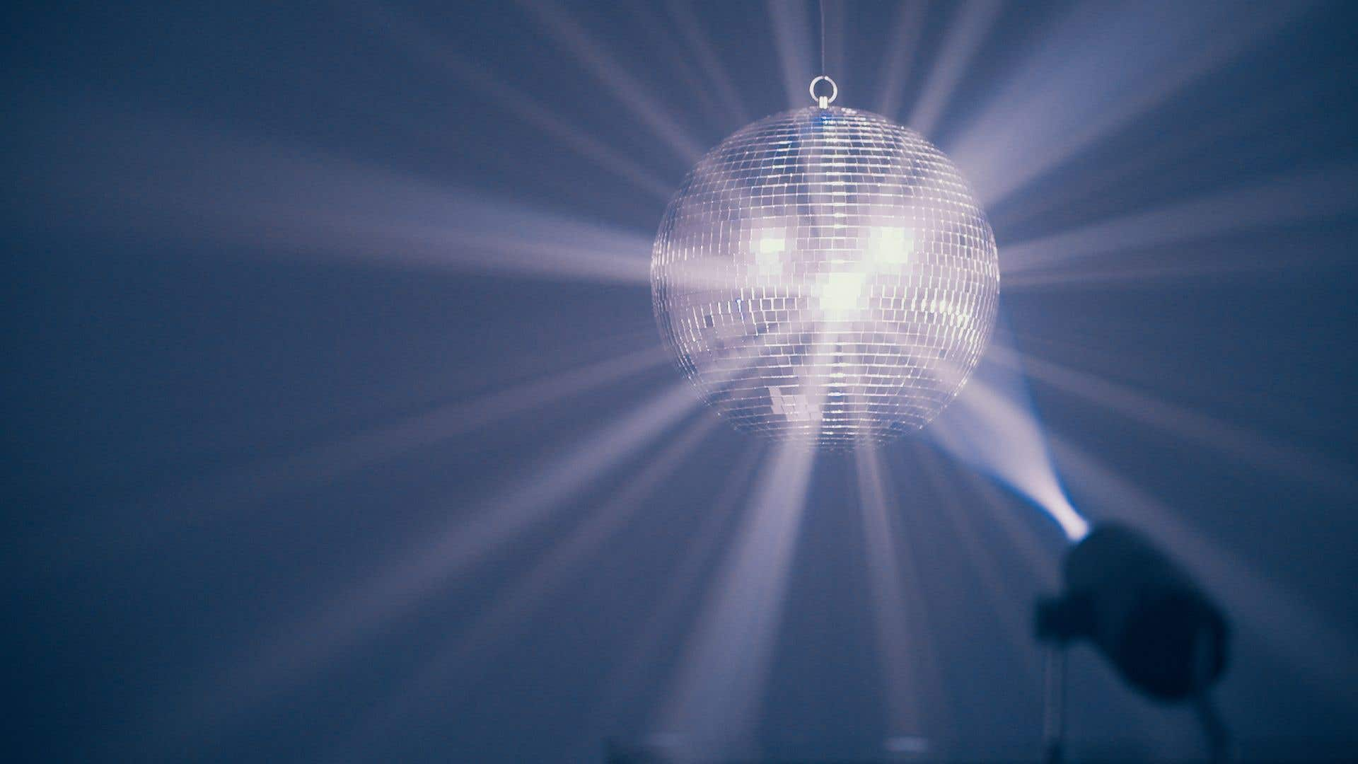 Una bola de discoteca cuelga, reflejando luz. Un foco brilla desde abajo, creando haces. Fondo azul oscuro.