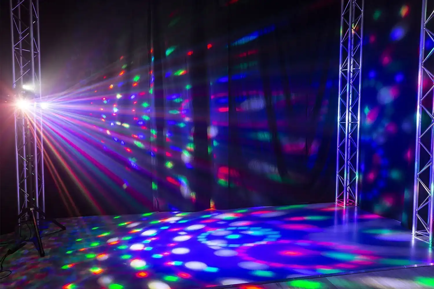 Un escenario con un foco que proyecta rayos coloridos. Las luces se reflejan en el suelo y el fondo.