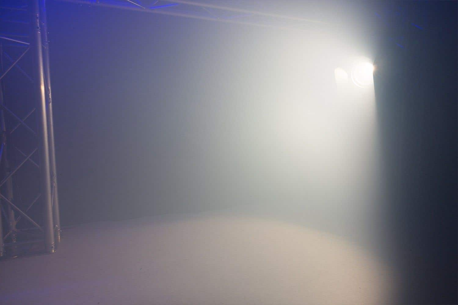 Una luz brillante de escenario ilumina una habitación llena de humo. Una estructura metálica está a la izquierda. La luz está enfocada, creando un ambiente nebuloso.