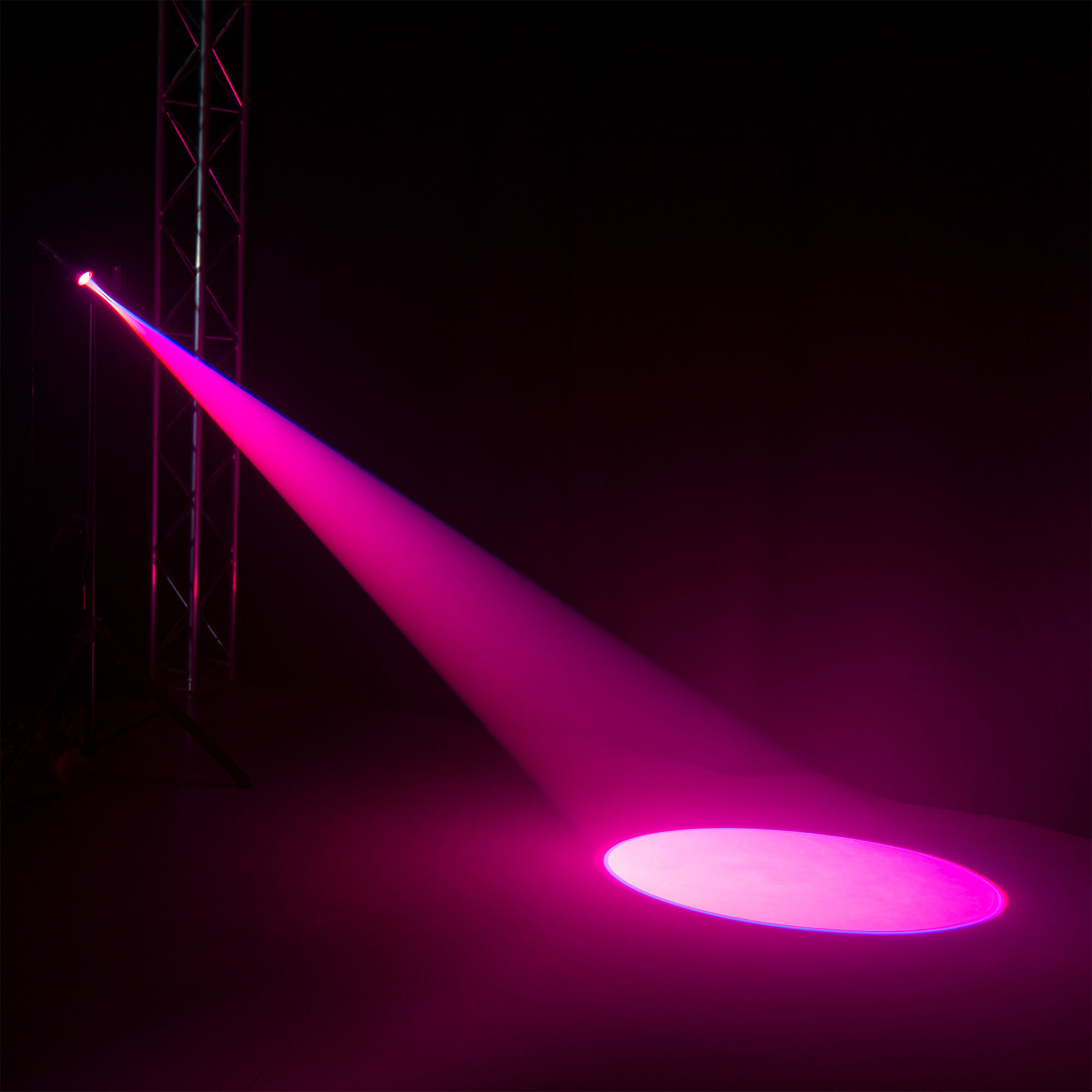 Un foco de escenario proyecta un haz rosa sobre el suelo. A la izquierda hay una estructura metálica, y el fondo es oscuro.