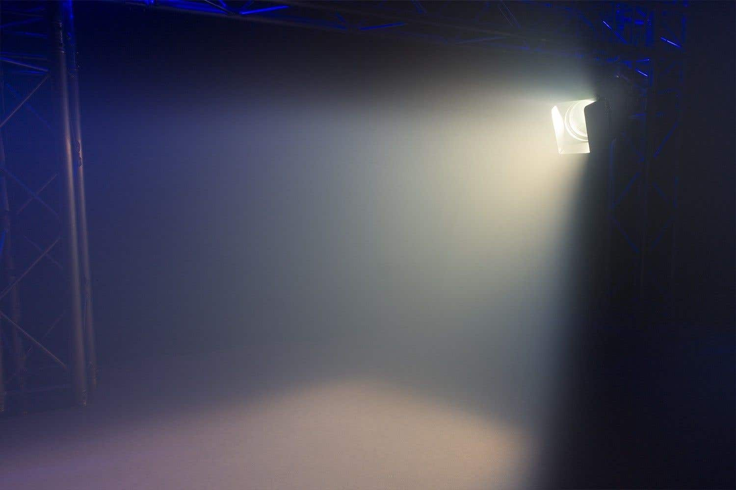 Un foco brilla en un escenario, creando un haz brillante contra un fondo azul oscuro y ahumado. Se ve el equipo del escenario.