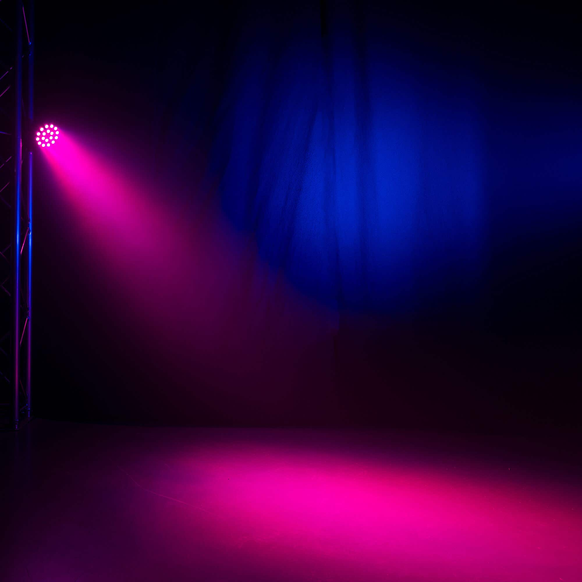 Un foco rosa ilumina un escenario oscuro. Una estructura metálica a la izquierda. Fondo azul y negro.